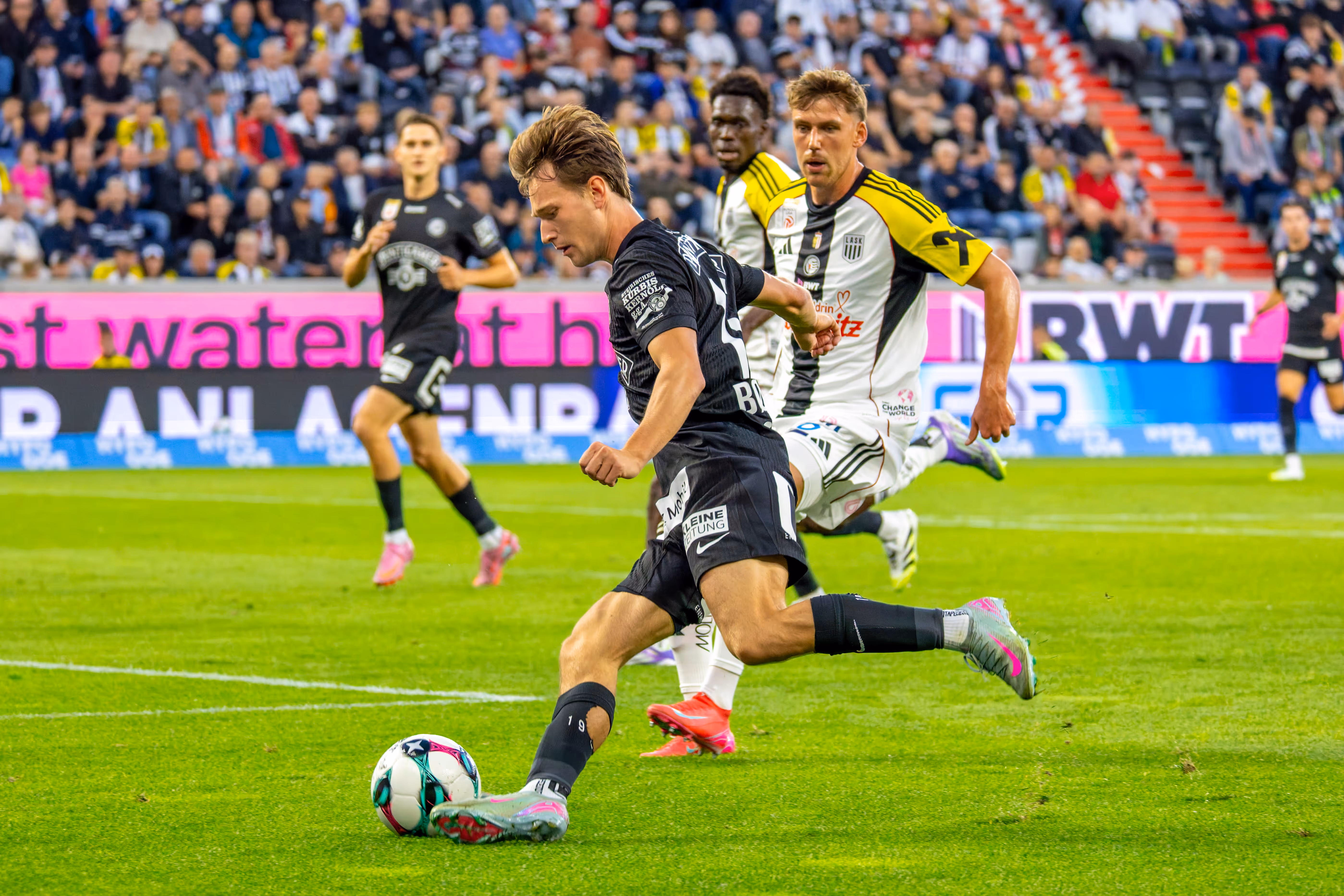 Ein Fußballspieler von Sturm Graz im schwarzen Trikot schießt den Ball während eines Bundesligaspiels, verfolgt von einem LASK-Spieler