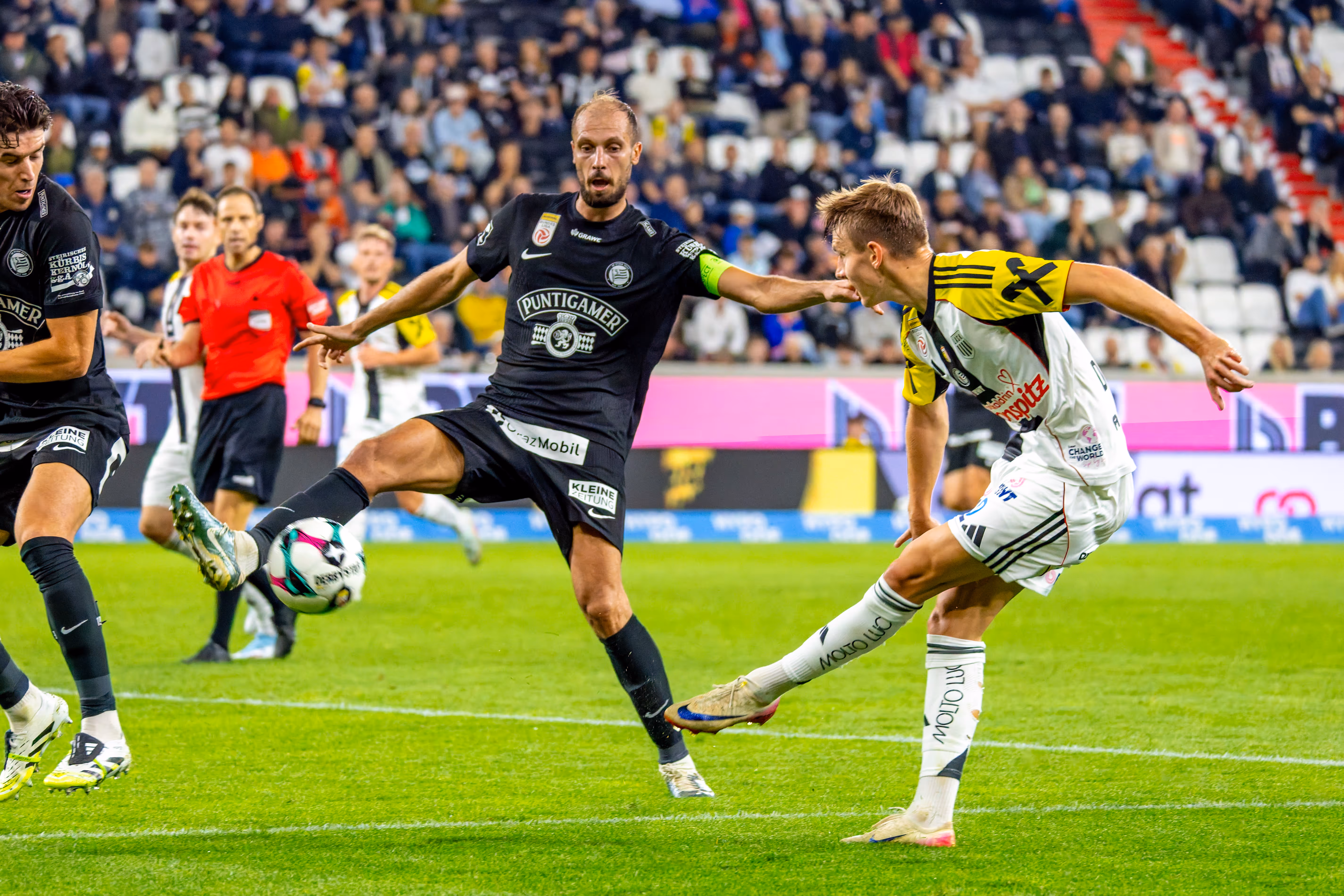 Ein Fußballspieler in dunklem Trikot schießt den Ball während eines Zweikampfs mit einem LASK-Spieler in hellem Trikot in einem Stadion
