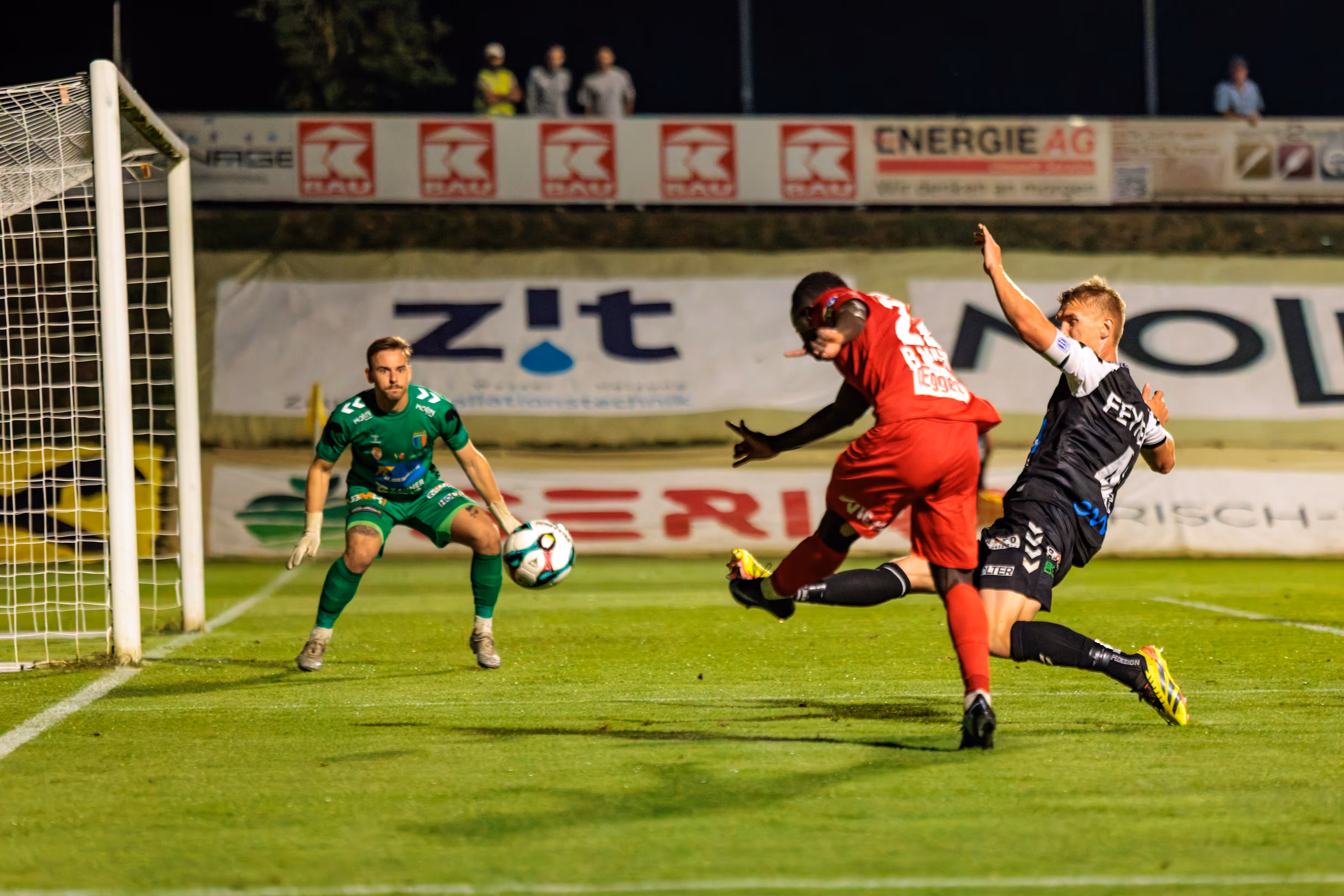 Ein Spieler des SKN St. Pölten in rotem Trikot schießt den Ball auf das Tor, während ein Welser Verteidiger in schwarz-weißem Trikot ihn zu blocken versucht und der Torwart zur Parade bereitsteht