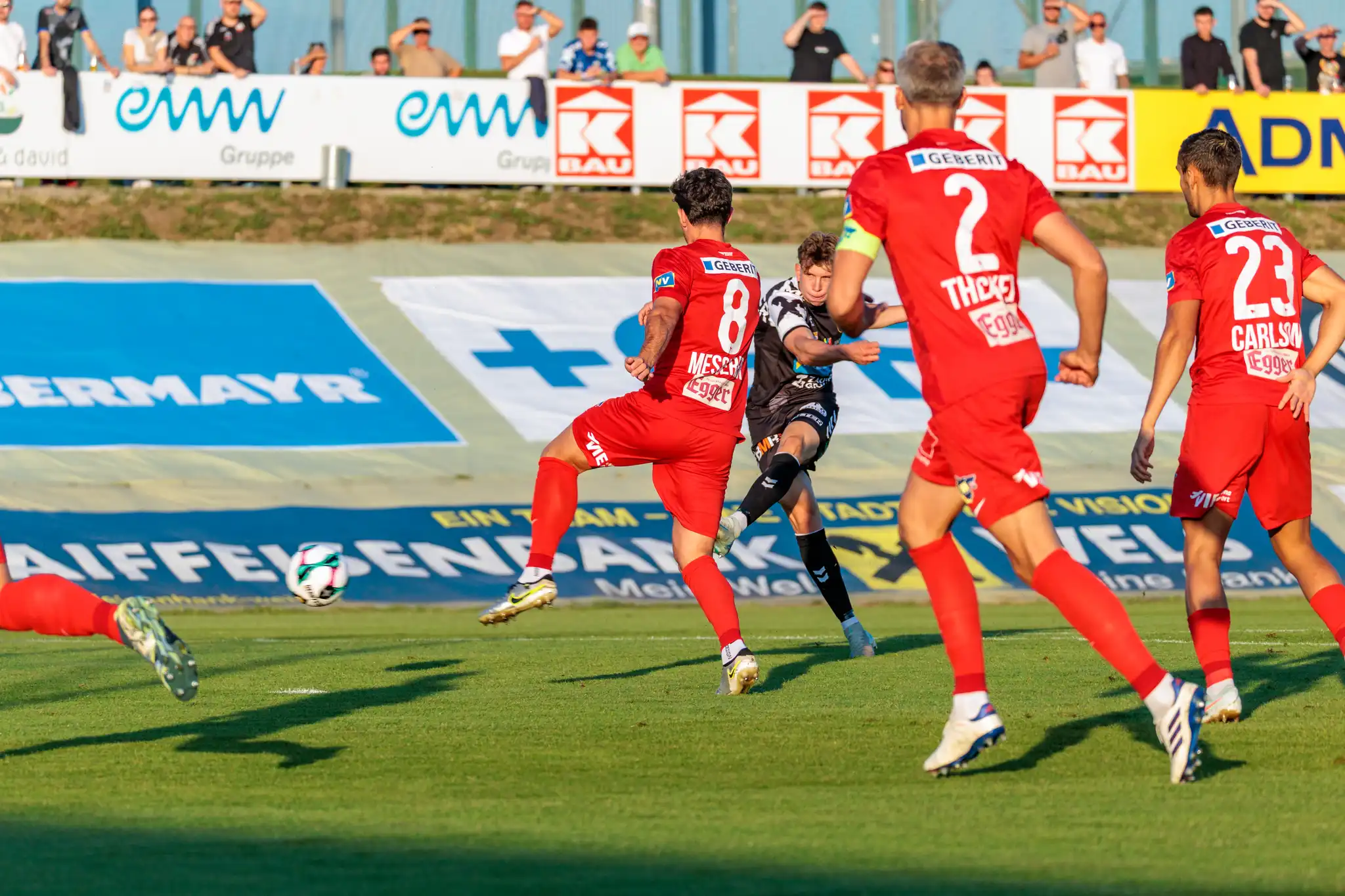 Ein Spieler der SPG HOGO Wels versucht im schwarz-weißen Trikot einen Schuss, der von mehreren Spielern des SKN St. Pölten in roten Trikots verdeckt wird
