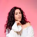 Porträt von Nicole Pecnik mit dunklem Lockenhaar und weißem Oberteil vor rosa Hintergrund