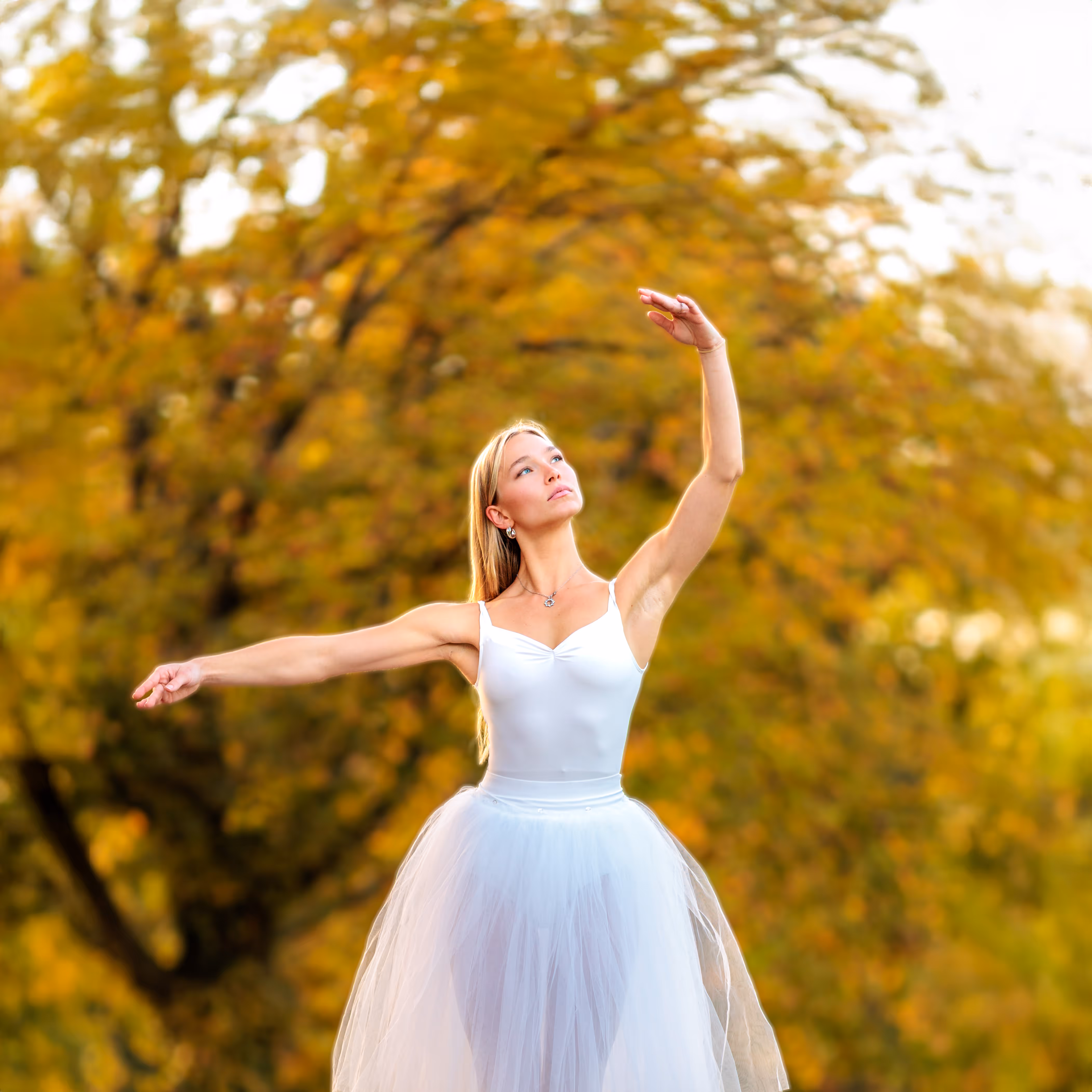 Eine anmutige Balletttänzerin in einem weißen Tutu posiert vor einem malerischen Hintergrund mit goldenem Herbstlaub