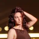 Eine junge Frau mit voluminösem, lockigem und rötlich-braunem Haar fährt sich mit der Hand durch die Haare und schaut nachdenklich nach oben