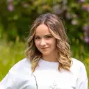 Eine junge Frau mit blonden Locken und einem weißen Pullover lächelt leicht in die Kamera vor einem natürlichen, unscharfen grünen Hintergrund  lens: RF70-200mm F2.8 L IS USM