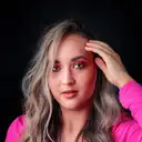 Nahaufnahme einer jungen Frau mit blond-braunem welligem Haar und auffälligem rosa Augen-Make-up, die mit der Hand an der Stirn in die Kamera blickt