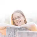 Eine junge Frau mit Brille lächelt und lehnt sich im goldenen Licht auf eine Betonmauer vor einem unscharfen Hintergrund  lens: 56mm F1.4 DC DN | Contemporary 018