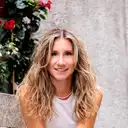 Nahaufnahme einer lächelnden Frau mit langen blonden Locken und Perlenkette vor einem leicht unscharfen Hintergrund aus einer strukturierten Wand und Pflanzen