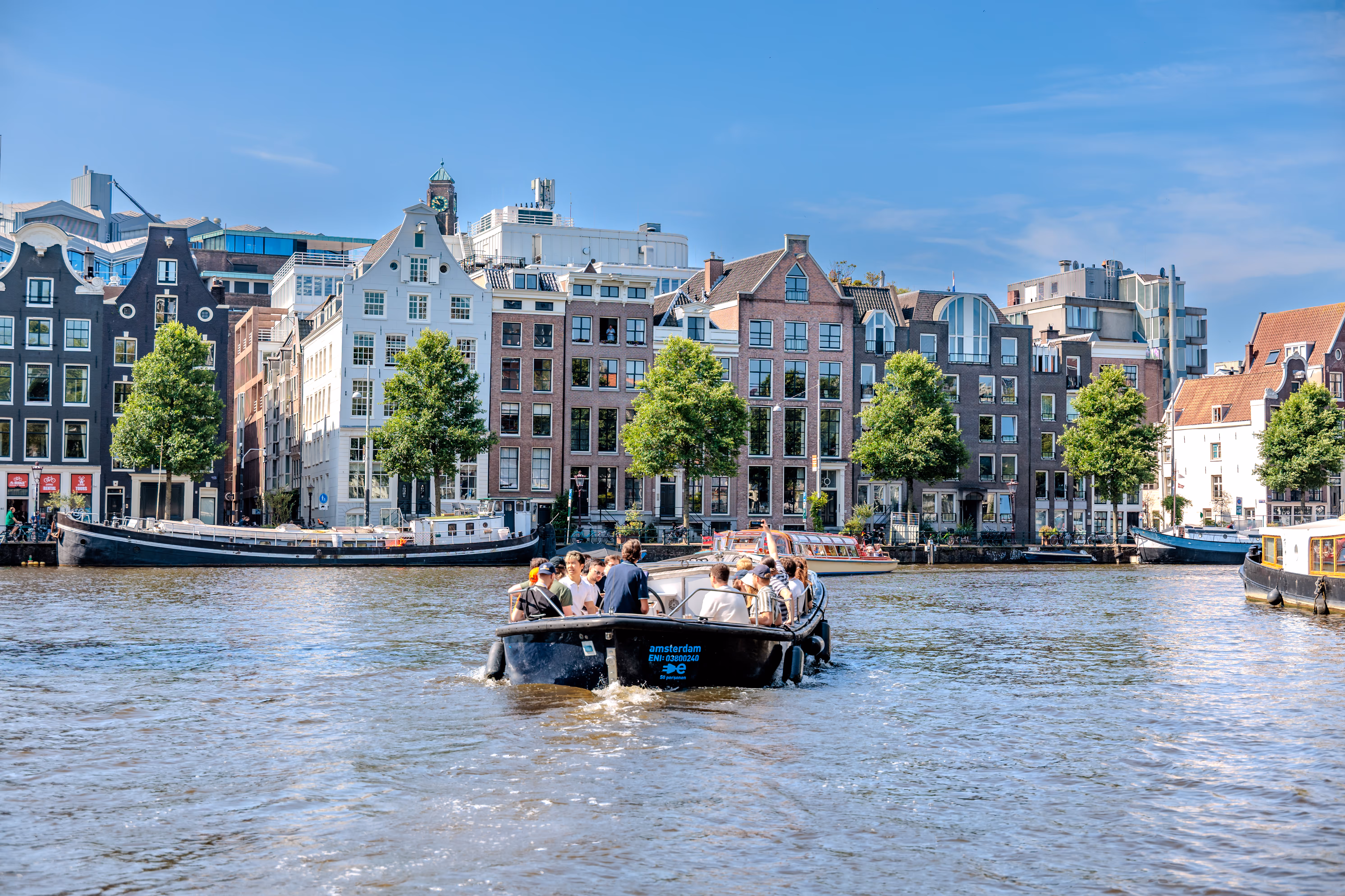 Ein Boot mit Passagieren fährt auf einem sonnigen Kanal in Amsterdam, umrahmt von den ikonischen Grachtenhäusern und Bäumen 