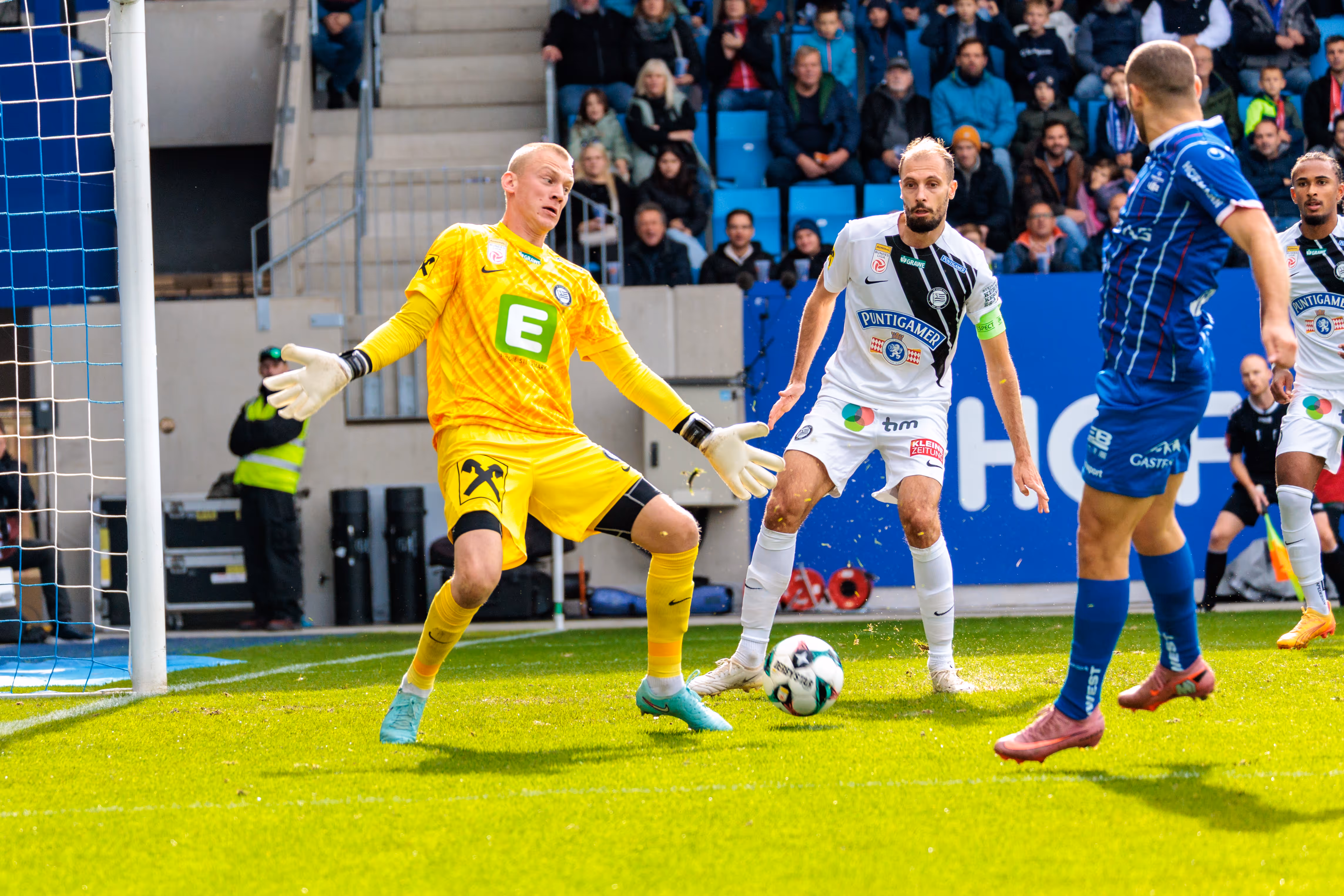 Sturm Graz-Torhüter Oliver Christensen in leuchtend gelber Tormannkleidung bei einer angespannten Abwehraktion im Strafraum gegen den FC Blau-Weiß Linz