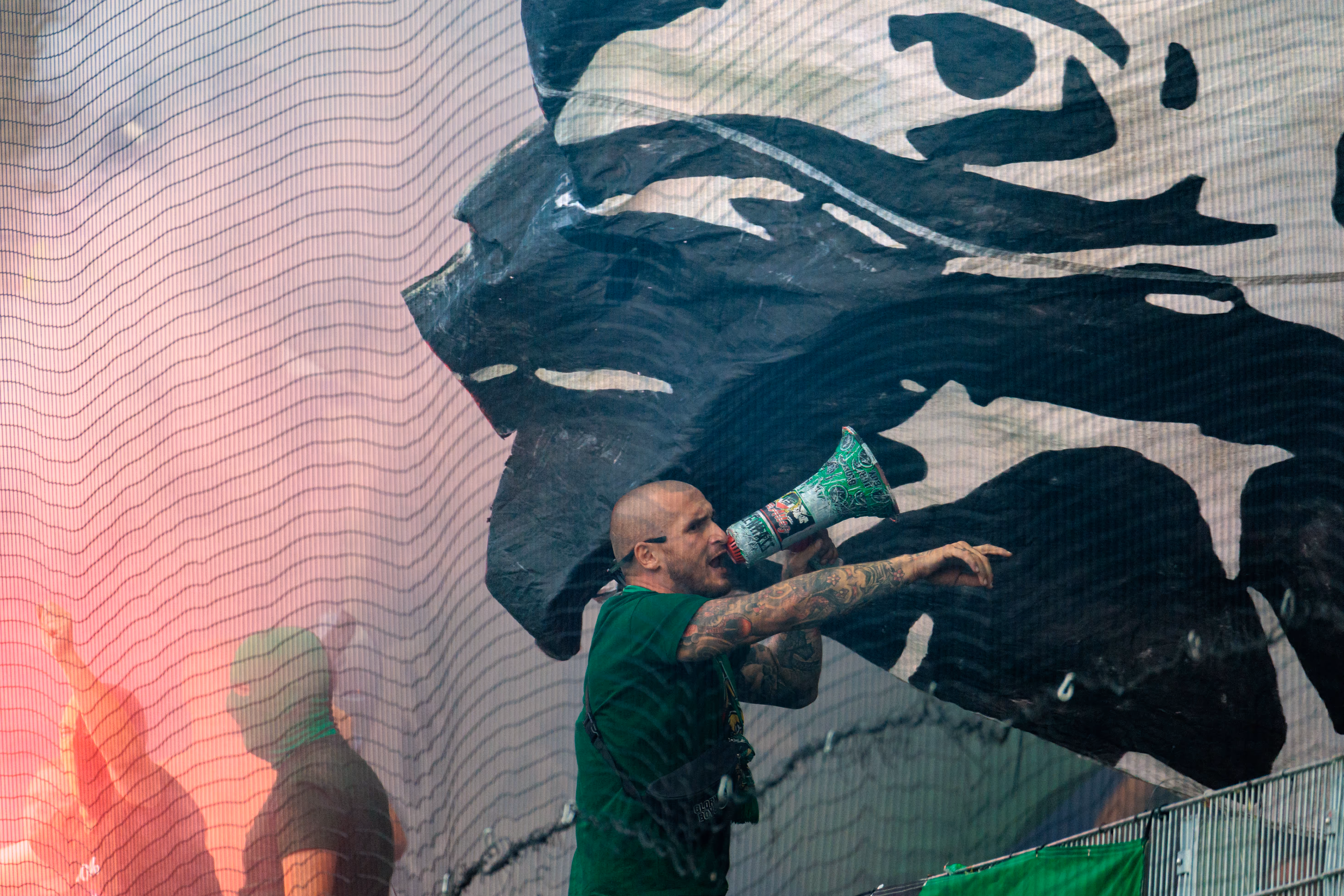 Ein SV Ried Fan mit kahlgeschorenem Kopf und tätowierten Armen brüllt mit einem grünen Megafon, umgeben von aufsteigendem rotem Pyrorauch hinter einem Sicherheitsnetz im Stadion