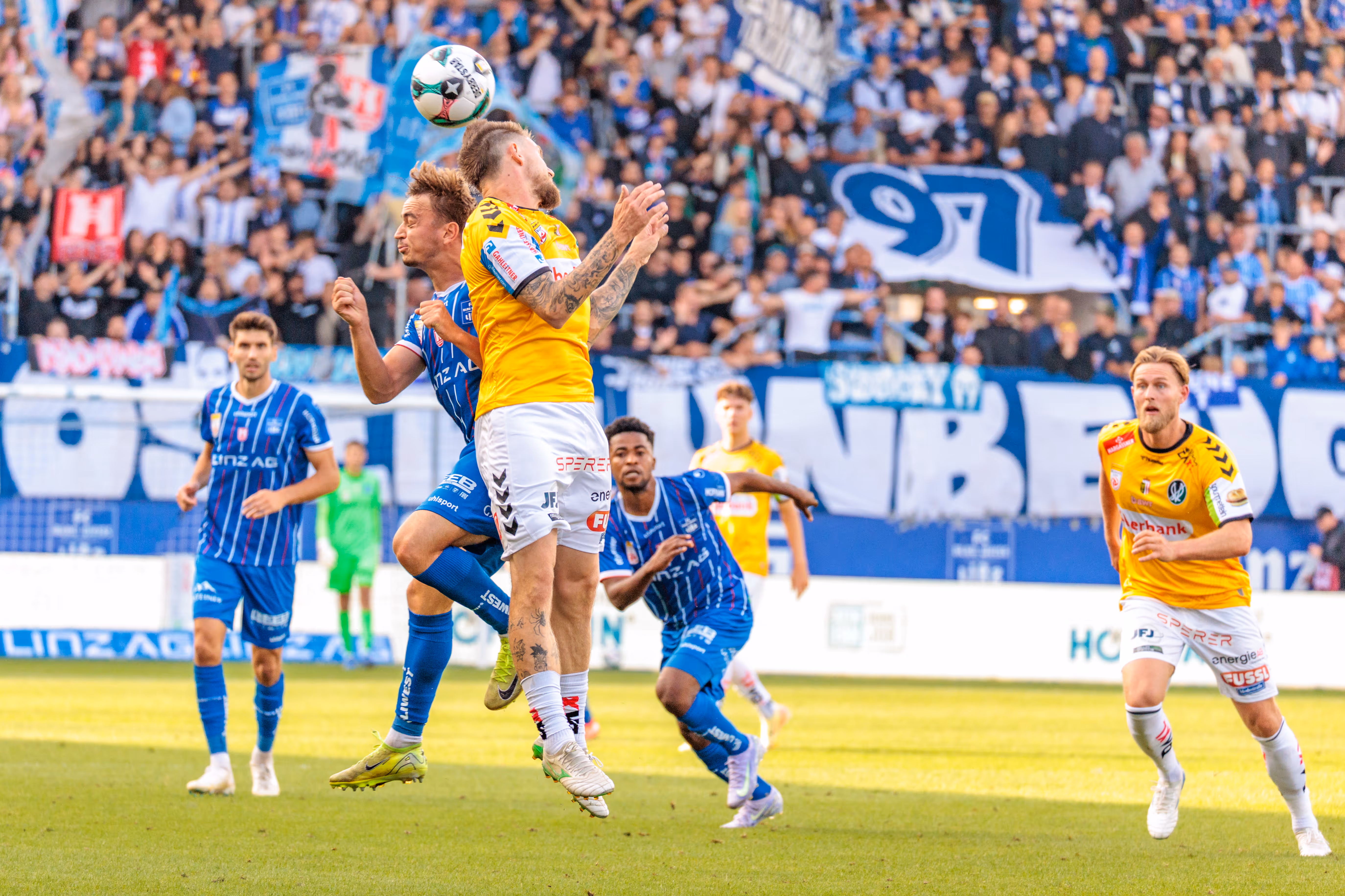 Ein Fußballspieler des SV Ried im gelben Trikot köpft den Ball während eines intensiven Duells mit einem Gegenspieler des FC Blau-Weiß Linz