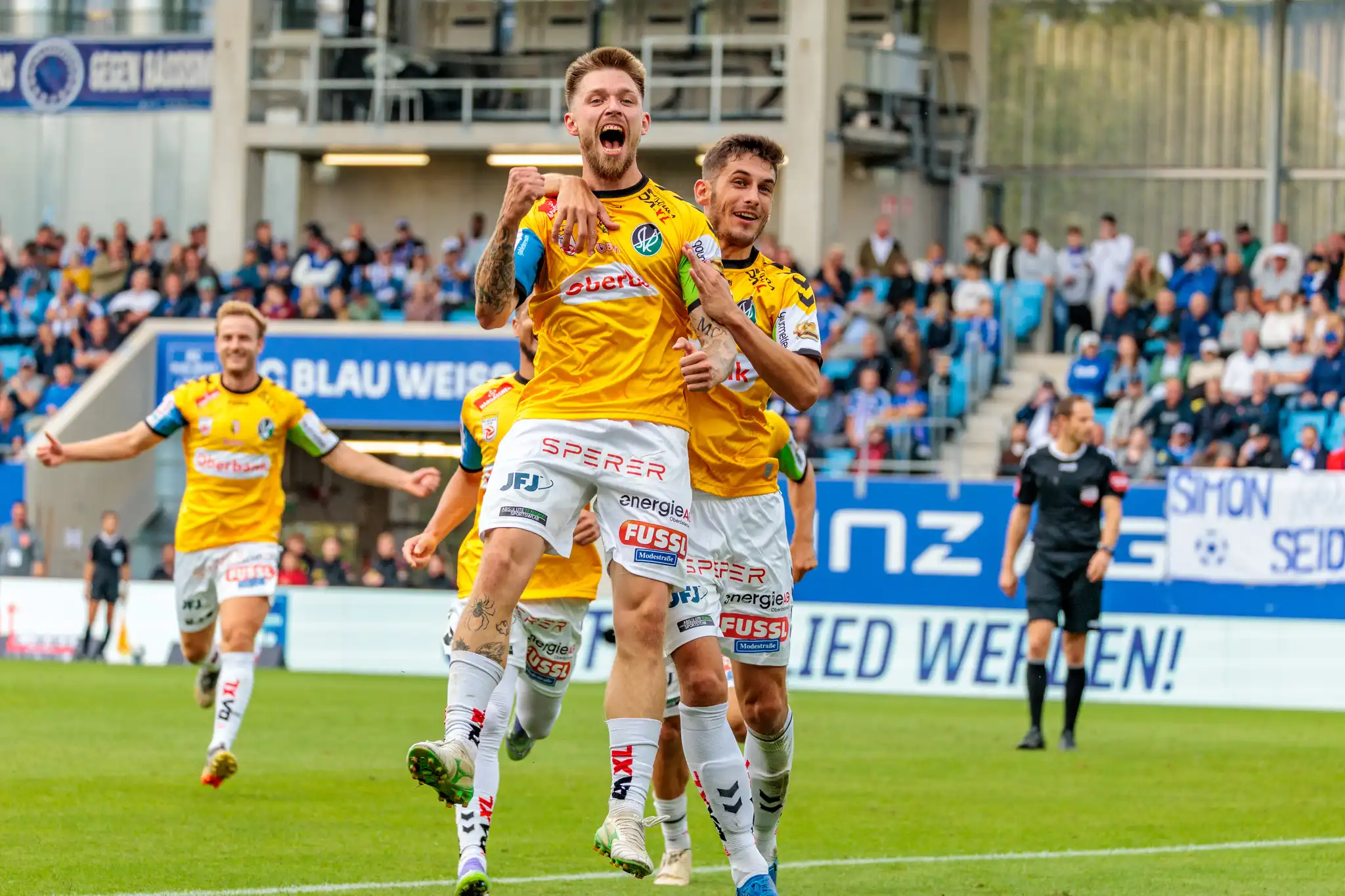 Zwei Fußballspieler des SV Ried in gelben Trikots jubeln ausgelassen gemeinsam nach einem erzielten Tor vor den Fans im Stadion