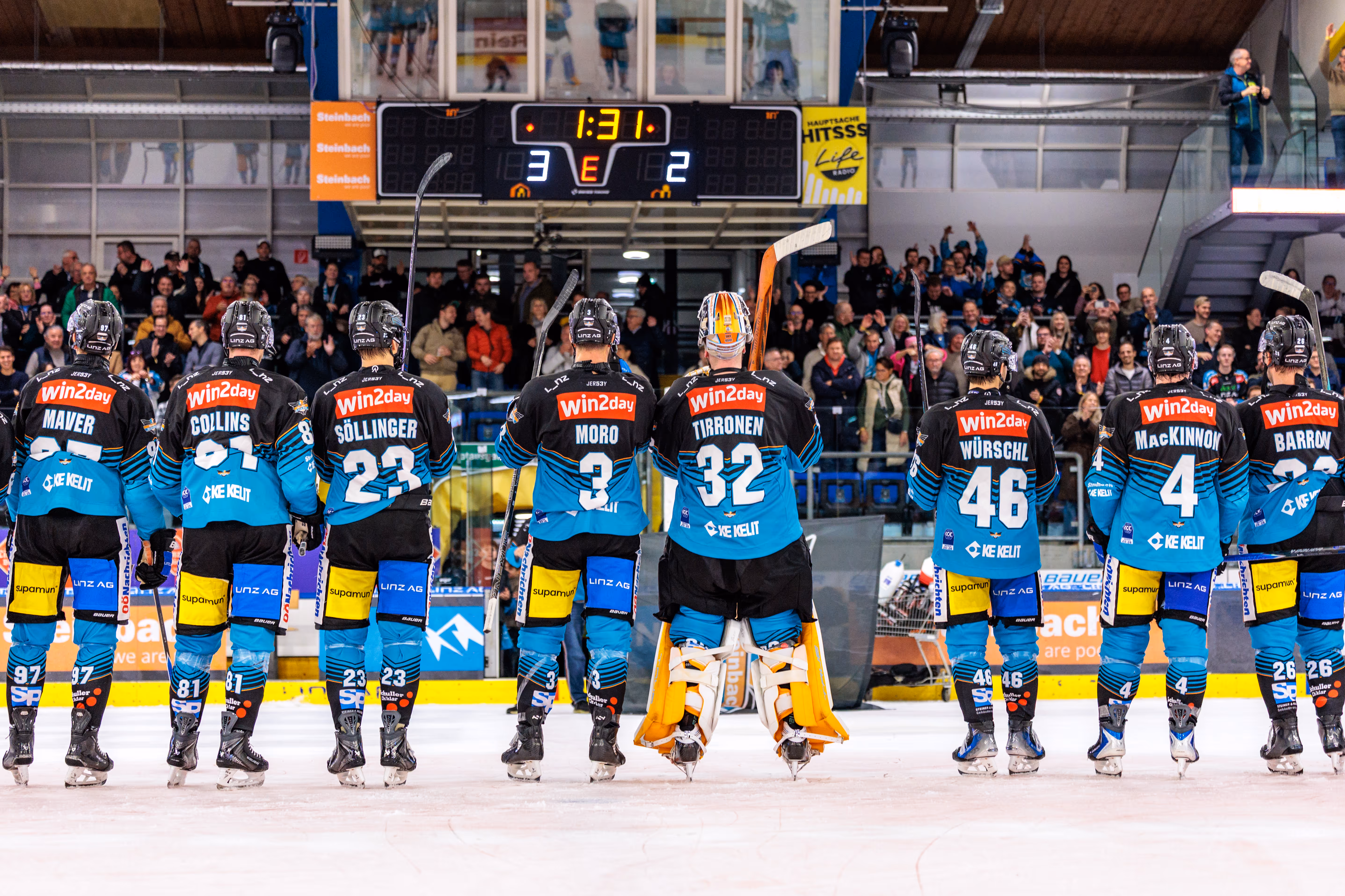 Die Eishockeymannschaft der Steinbach Black Wings Linz steht von hinten fotografiert in einer Reihe auf dem Eis vor der Anzeigetafel, die den 3:2-Sieg anzeigt