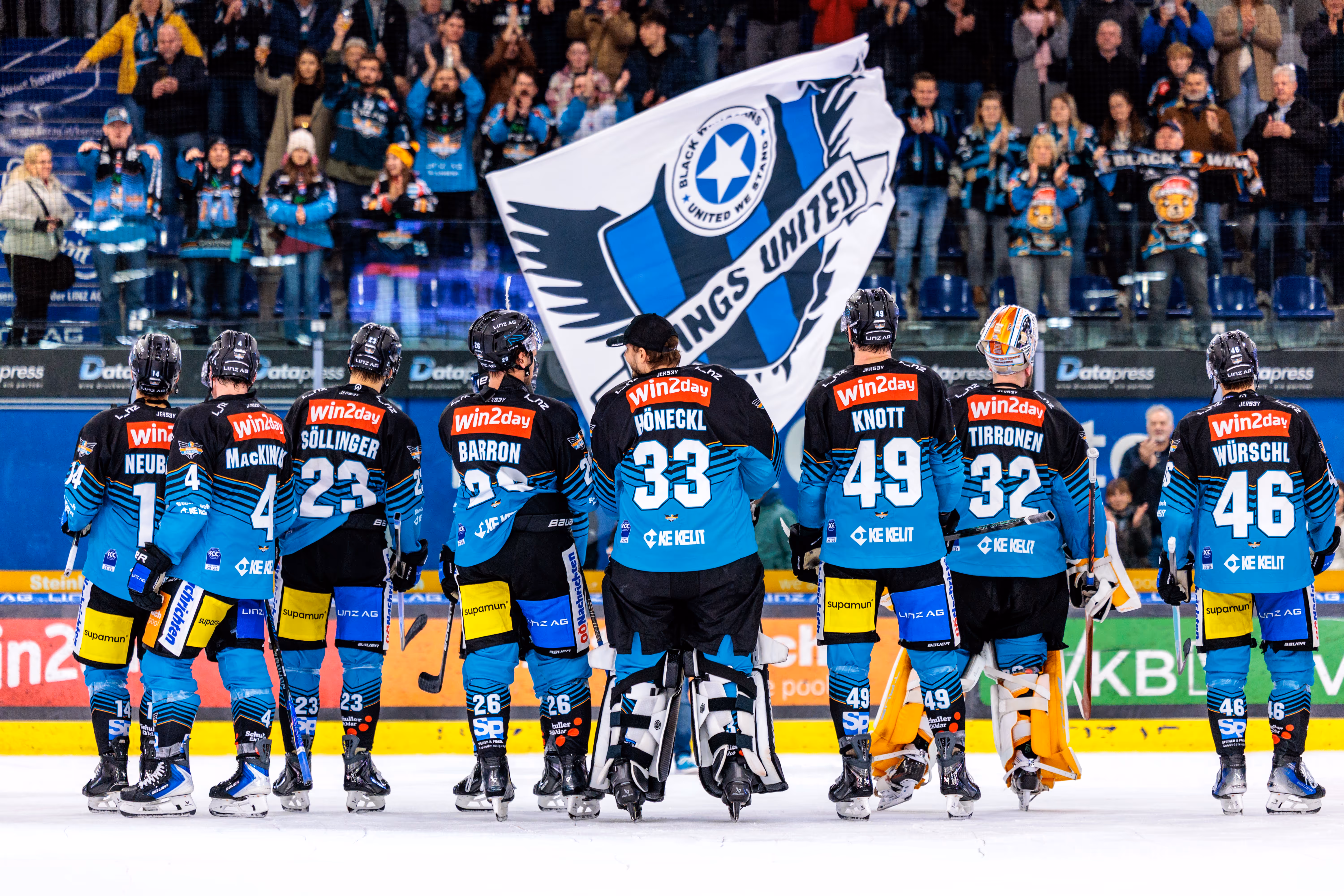 Die Eishockey-Spieler der Black Wings Linz stehen von hinten auf dem Eis vor einer großen Fanflagge und applaudierenden Zuschauern