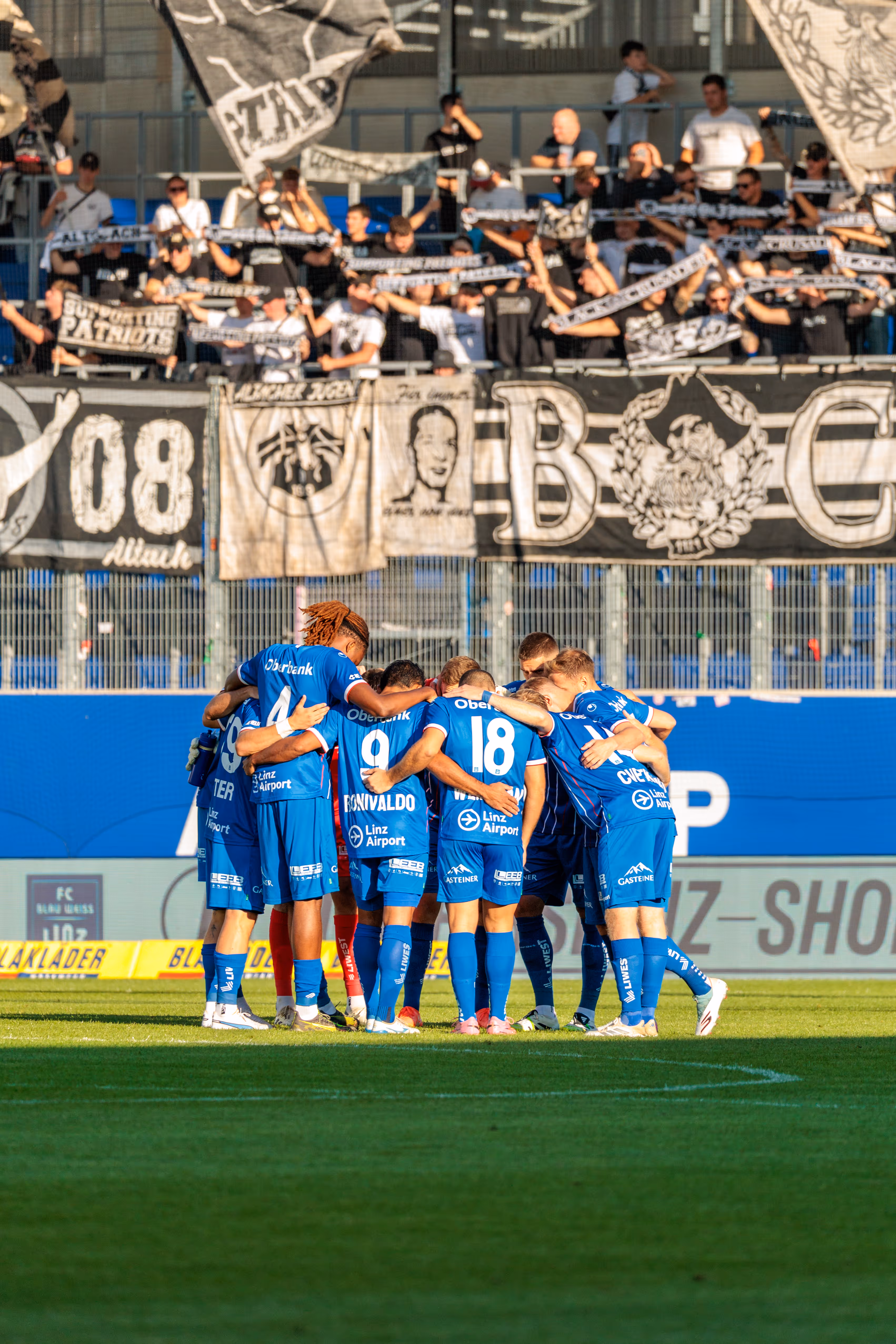 Die Fußballmannschaft des FC Blau-Weiß Linz bildet einen engen Kreis zur Teambesprechung vor der Fankurve mit den gegnerischen Zuschauern