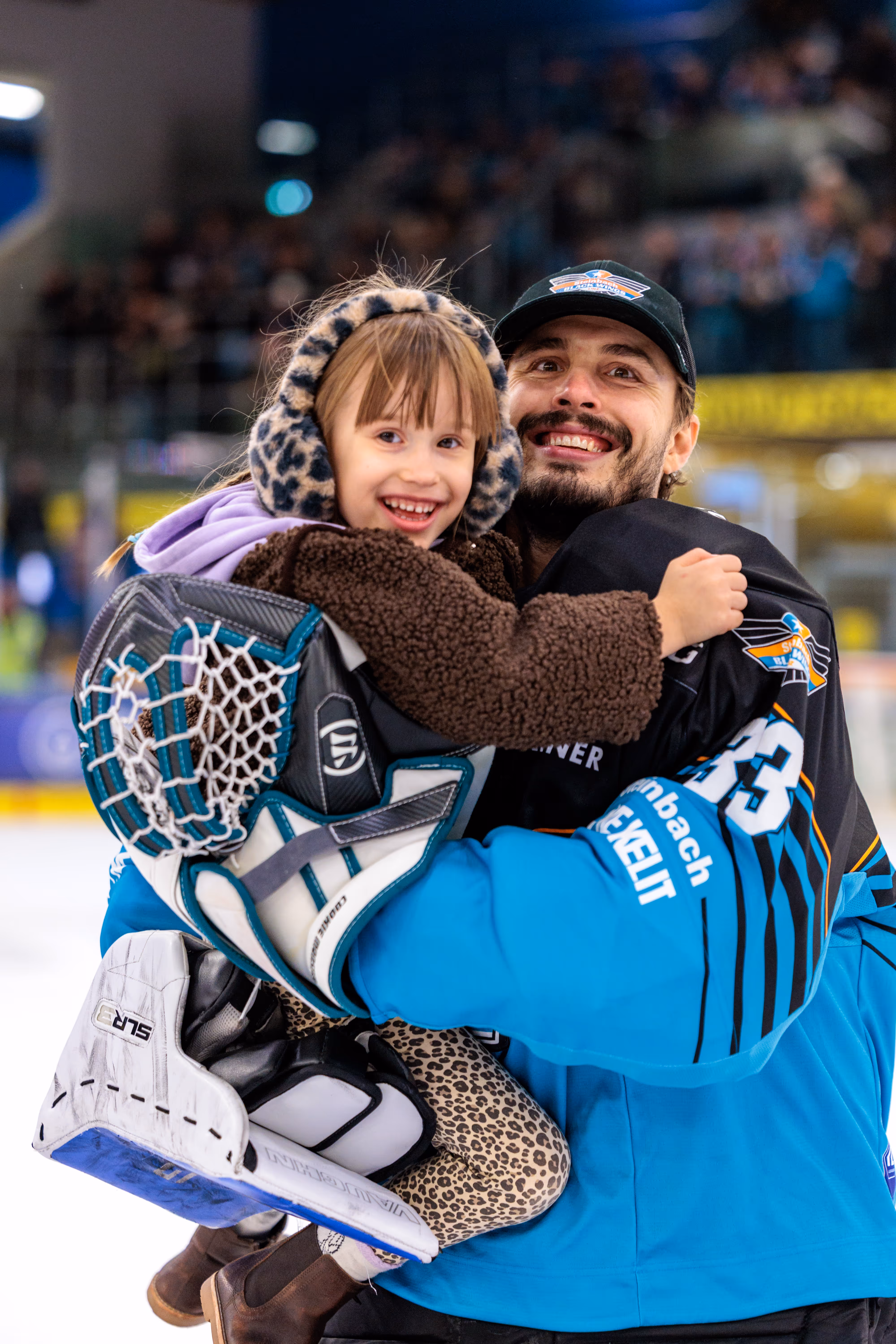 Der Eishockey-Torhüter Thomas Höneckl der Black Wings Linz mit der Trikotnummer 33 lächelt, während er ein fröhliches Kind in der Eisarena umarmt