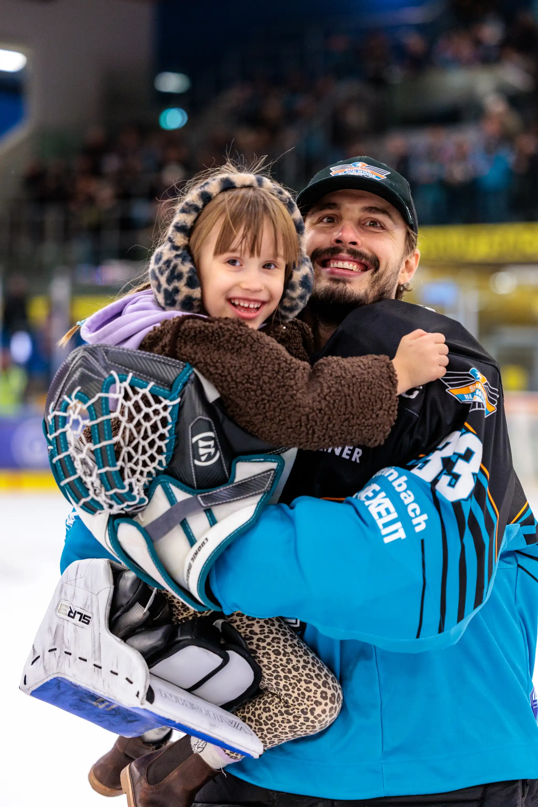 Der Eishockey-Torhüter Thomas Höneckl der Black Wings Linz mit der Trikotnummer 33 lächelt, während er ein fröhliches Kind in der Eisarena umarmt