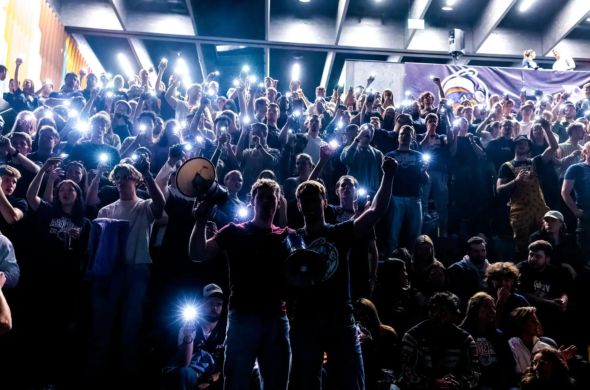Eine große, jubelnde Menschenmenge leuchtet bei einem Basketball-Halbfinale mit Handy-Taschenlampen in einer dunklen Kepler Hall an der JKU in Linz