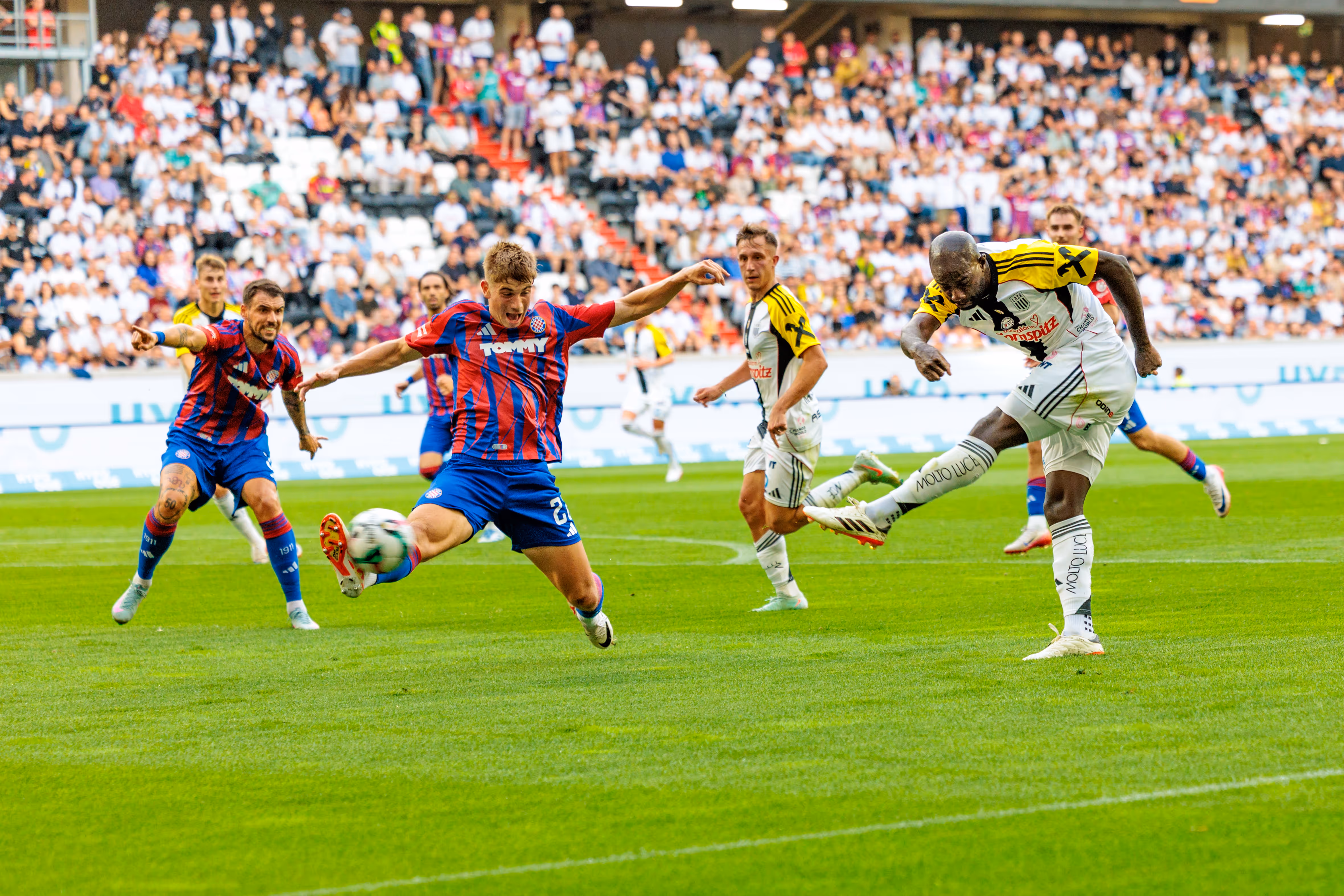 Ein Fußballspieler des LASK schießt dynamisch den Ball während eines Spiels im Stadion, umgeben von gegnerischen Spielern und Zuschauern