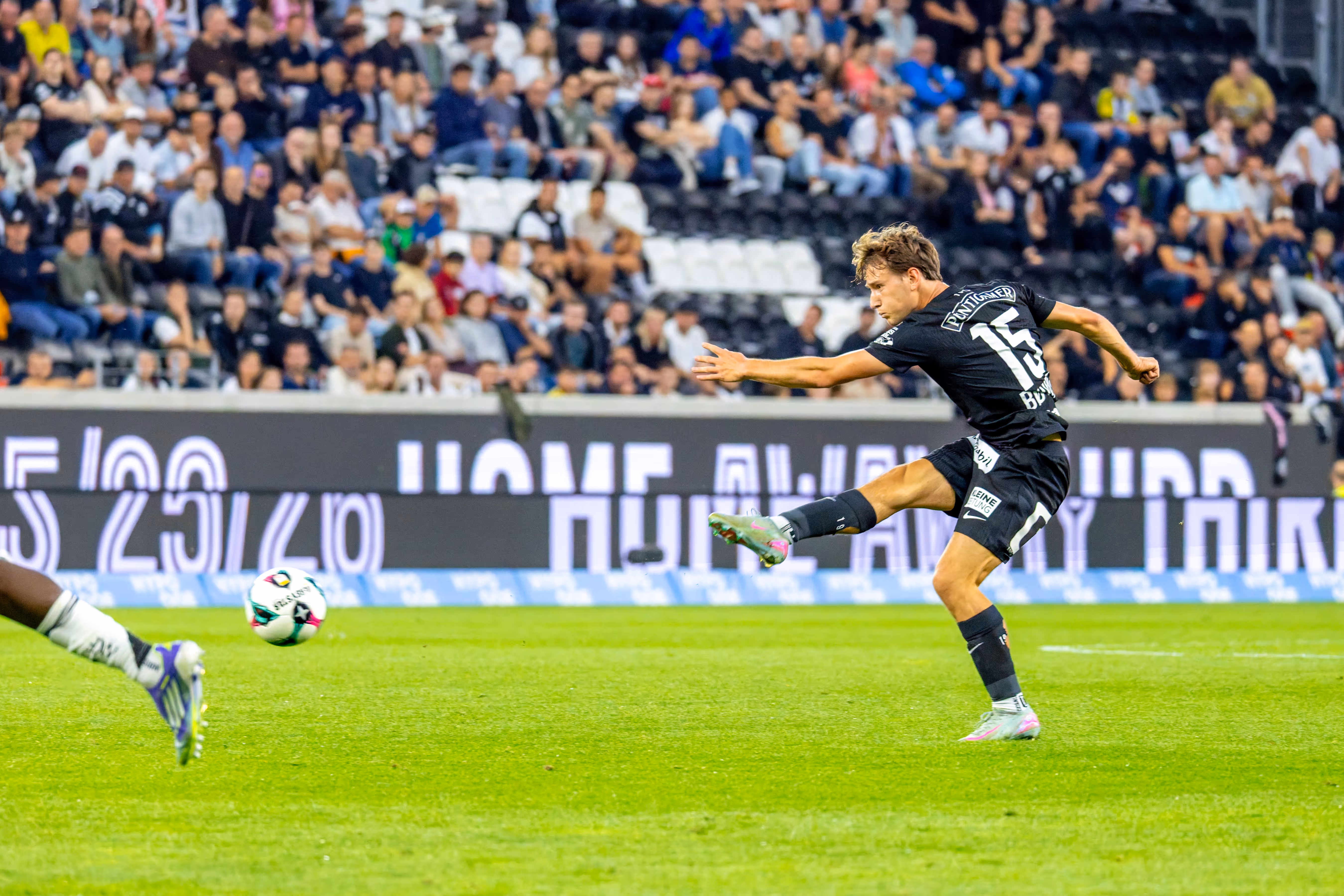 Ein Fußballspieler in schwarzem Trikot mit der Nummer 15 ist in voller Schussbewegung vor unscharfen Zuschauern