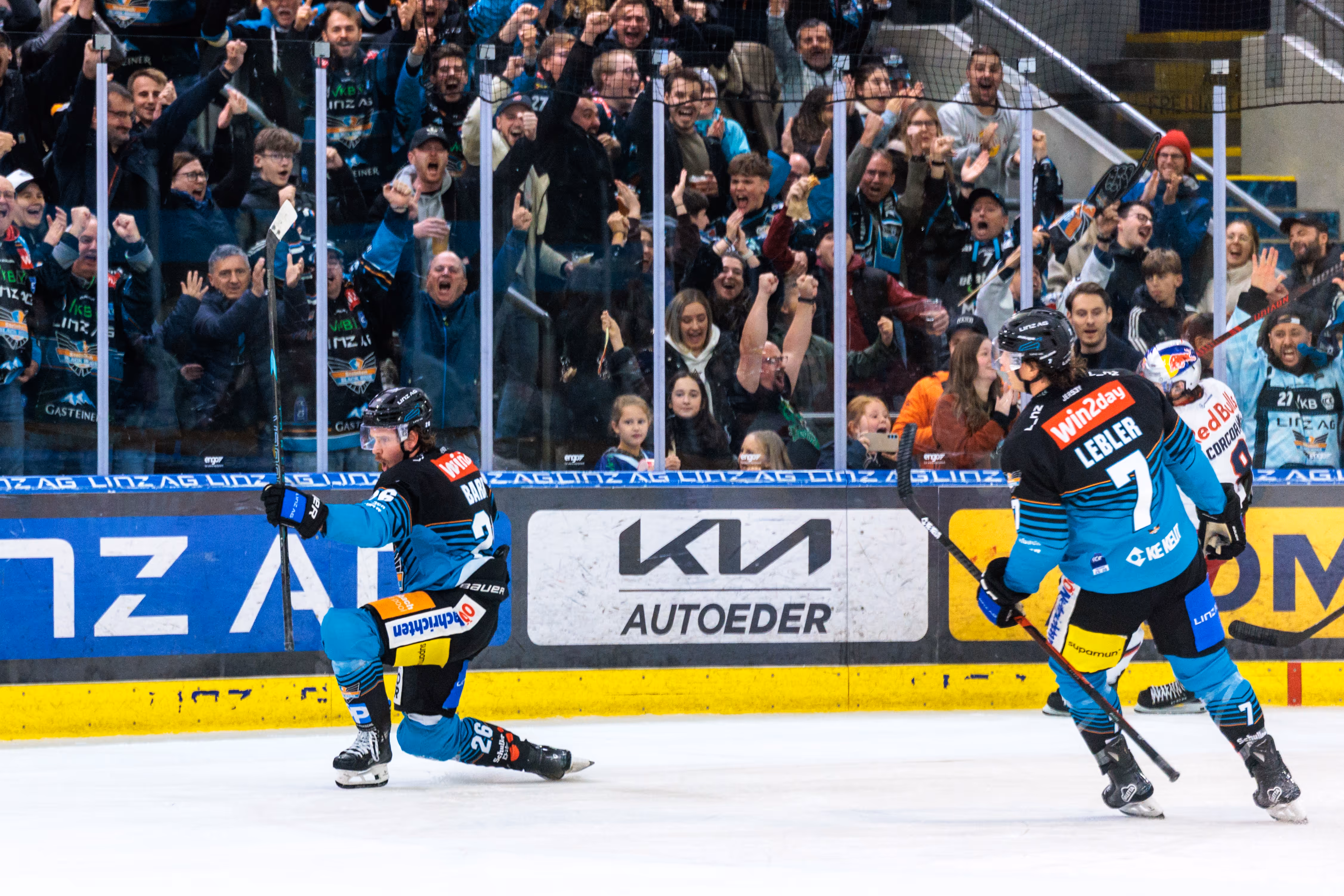 Der Eishockeyspieler Travis Barron (#26) der Steinbach Black Wings Linz feiert kniend auf dem Eis ein Tor, während die Fans und Kapitän Brian Lebler (#7) im Hintergrund jubeln