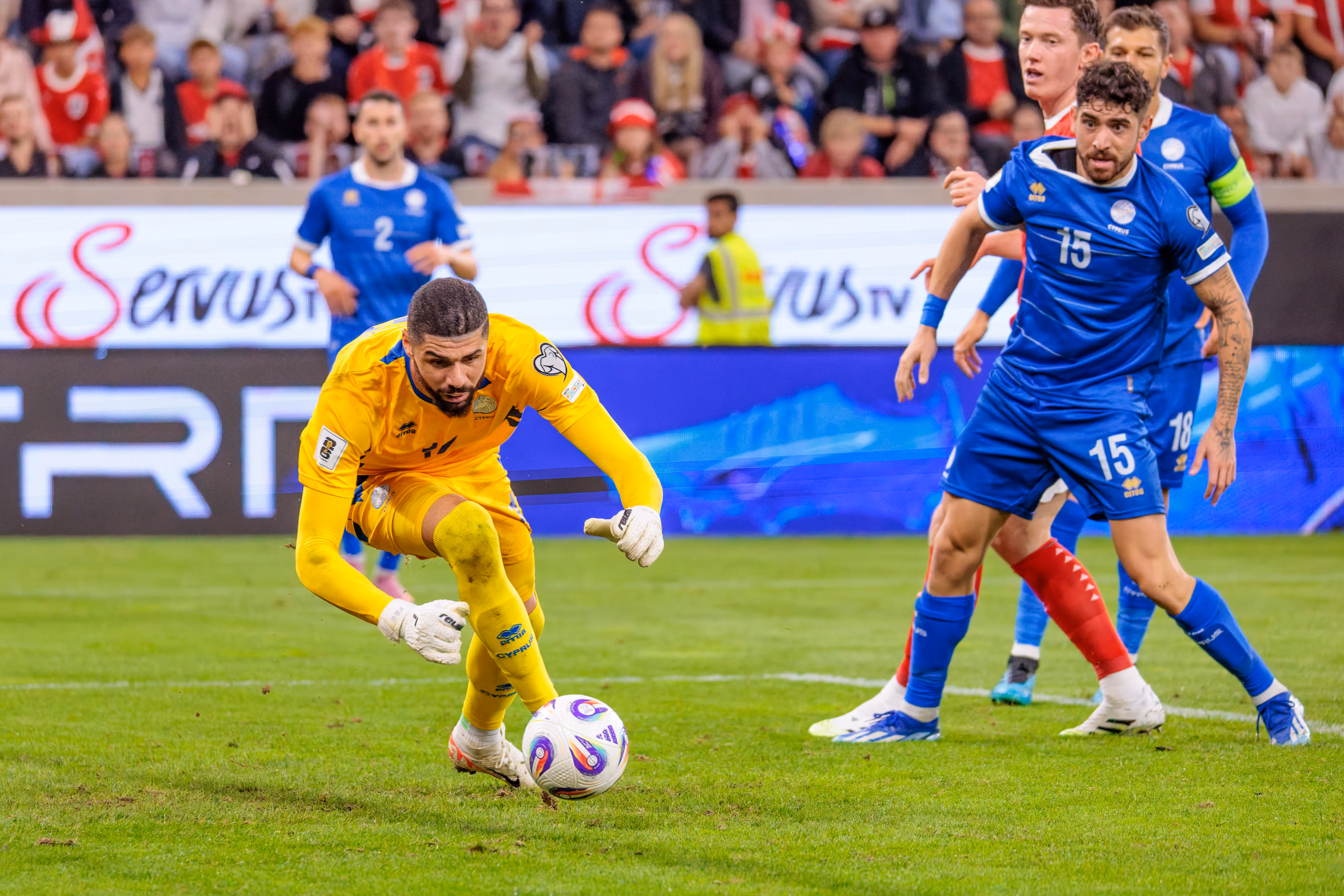 Der zypriotische Torhüter Fabiano im gelben Trikot kontrolliert den Ball am Boden, während seine Teamkollegen Christos Shelis (Nummer 15) und Kostas Pileas (Nummer 2) im Hintergrund positioniert sind