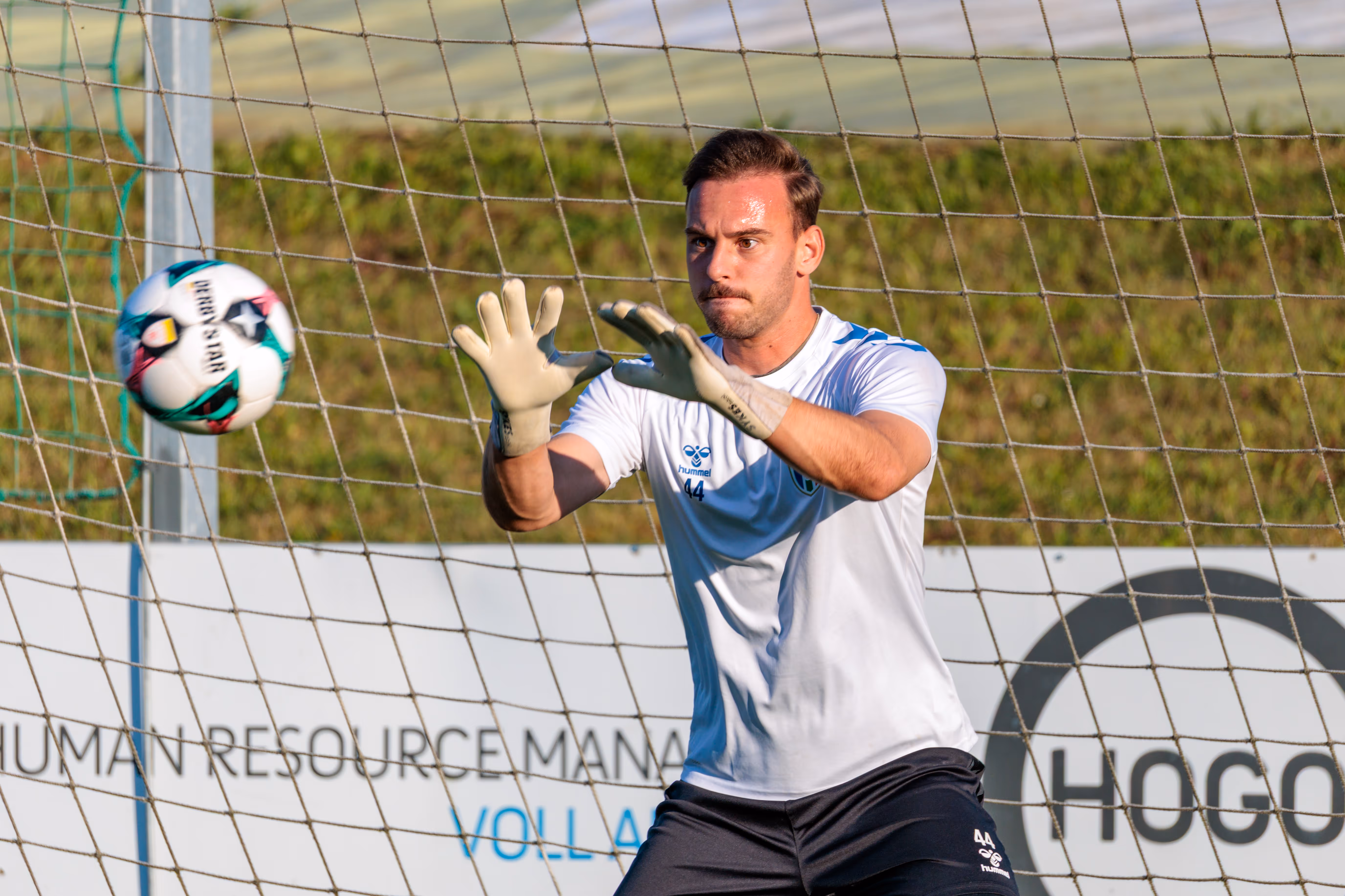Der Torwart der SPG HOGO Wels im weißen Trikot fängt konzentriert einen Fußball beim Aufwärmen vor dem Tornetz