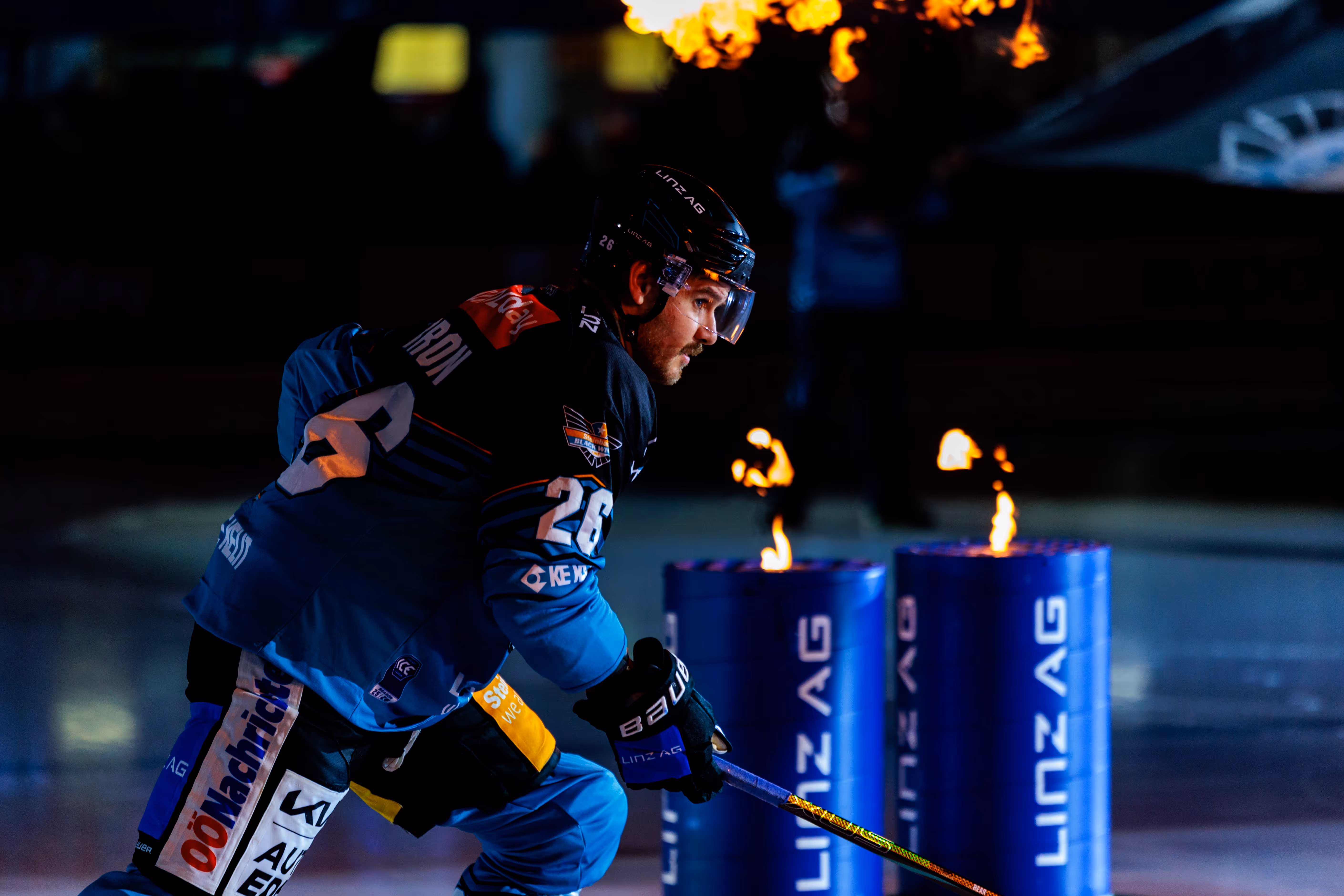 Eishockeyspieler Travis Barron in blauer und schwarzer Uniform gleitet beim Einlauf auf dem Eis, während Flammen aus Feuersäulen schlagen
