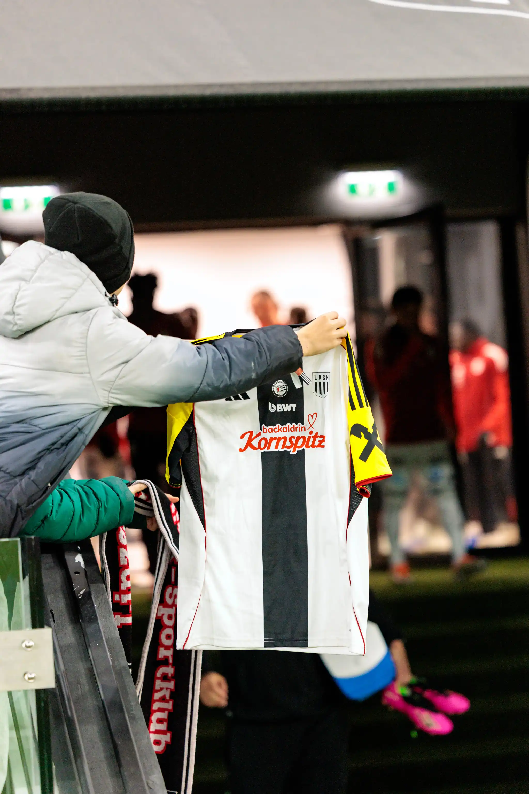 Ein LASK-Fan in grauer Jacke und schwarzer Mütze hält ein weiß-schwarz gestreiftes Fußballtrikot des LASK mit den Sponsoren BWT und Kornspitz an einem Geländer im Stadion hoch