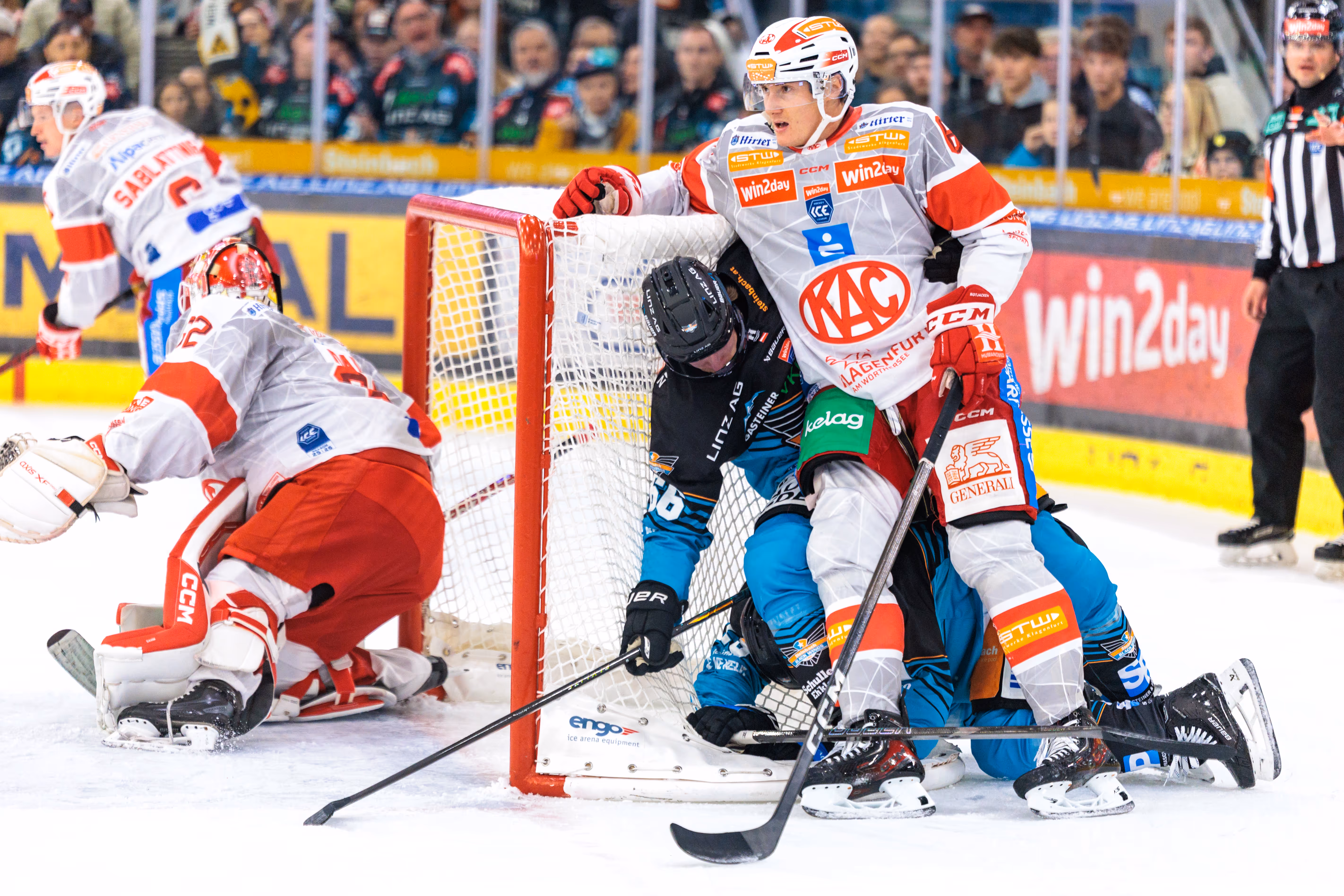 Eine tumultartige Szene im Eishockey-Spiel zwischen dem EC-KAC und den Black Wings Linz, bei der mehrere Spieler direkt vor dem Tor aneinandergeraten. Dabei stützt sich ein Spieler des KAC entspannt am eigenen Tor ab, während zwei Spieler der Black Wings am Tornetz liegen