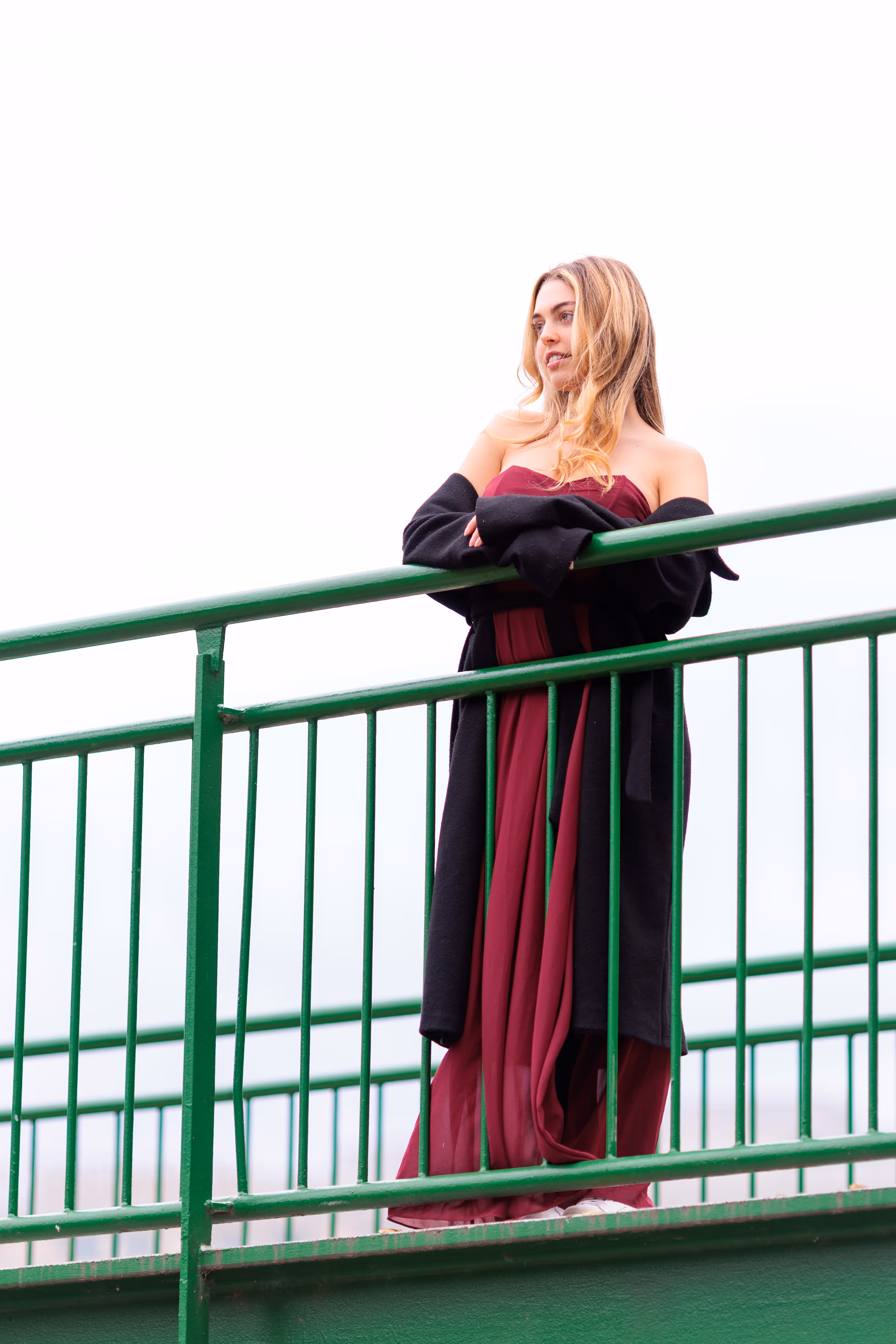 Das Model Valentina lehnt in einem eleganten roten Kleid und einem schwarzen Mantel lächelnd am grünen Geländer einer Brücke im Wiener Stadtpark