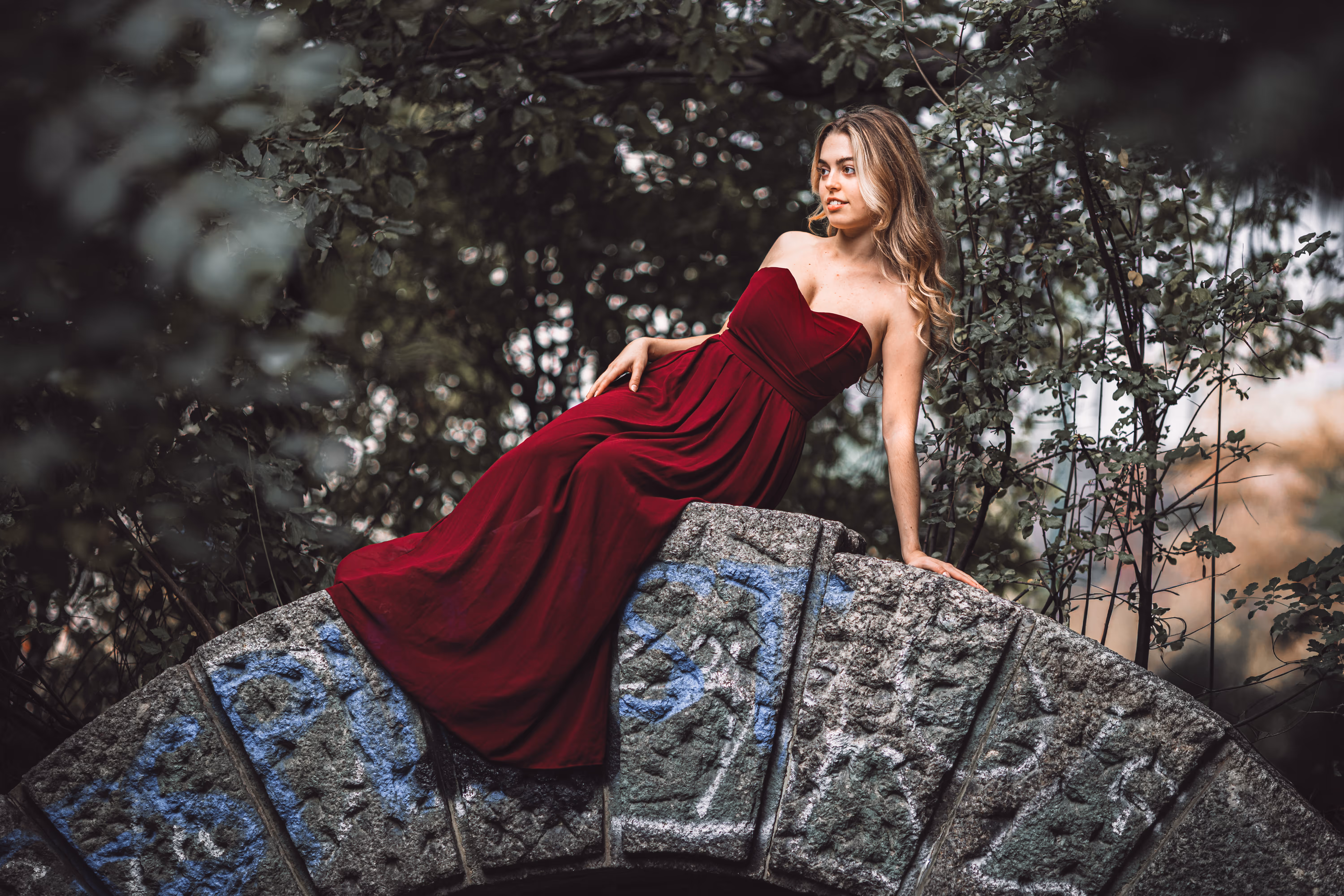 Model Valentina posiert elegant in einem langen, roten Kleid auf dem Bogen einer verwitterten Steinbrücke im Wiener Stadtpark