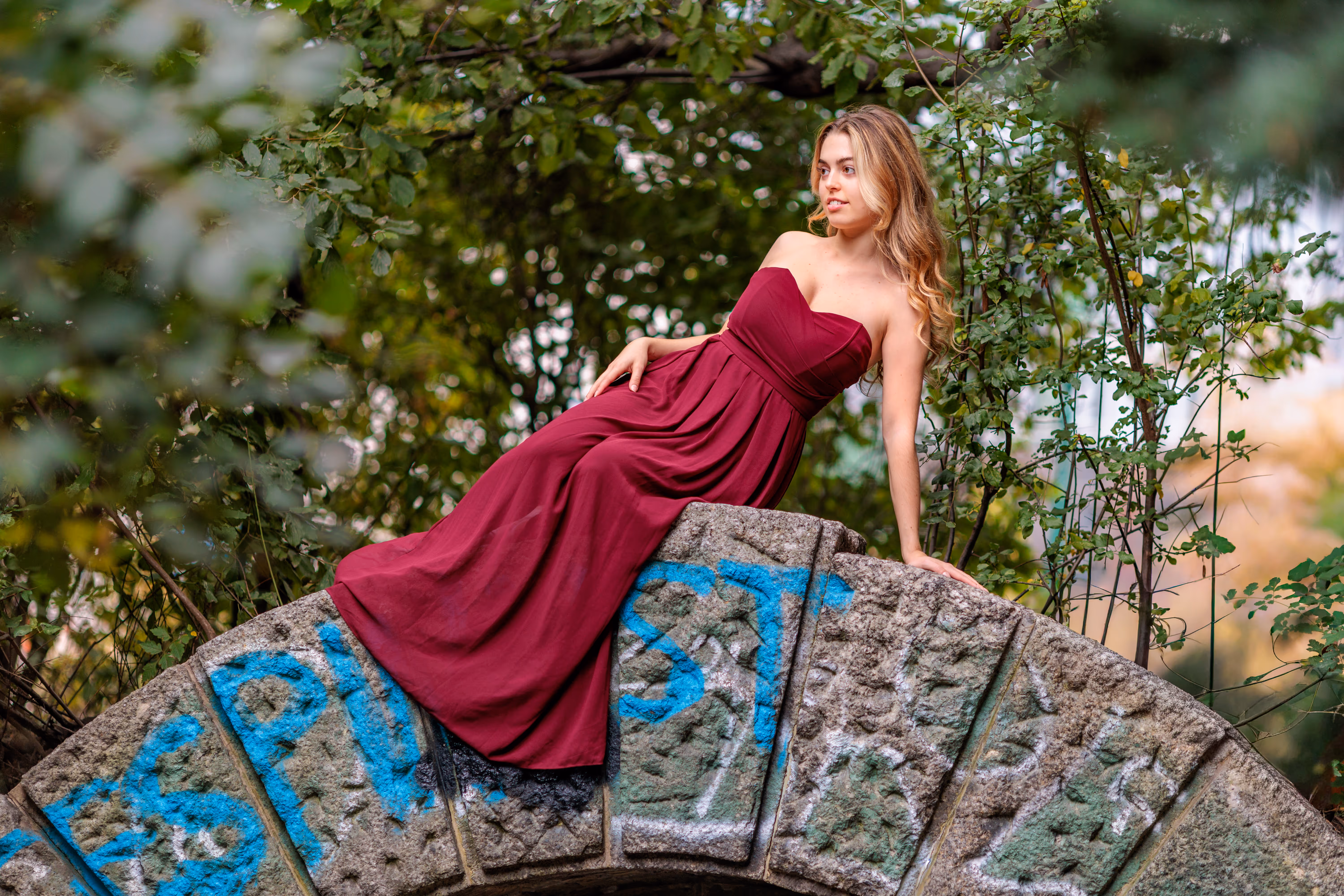 Model Valentina posiert während ihres Fotoshootings im Stadtpark elegant in einem roten Abendkleid auf dem Bogen einer alten Steinbrücke mit Graffiti