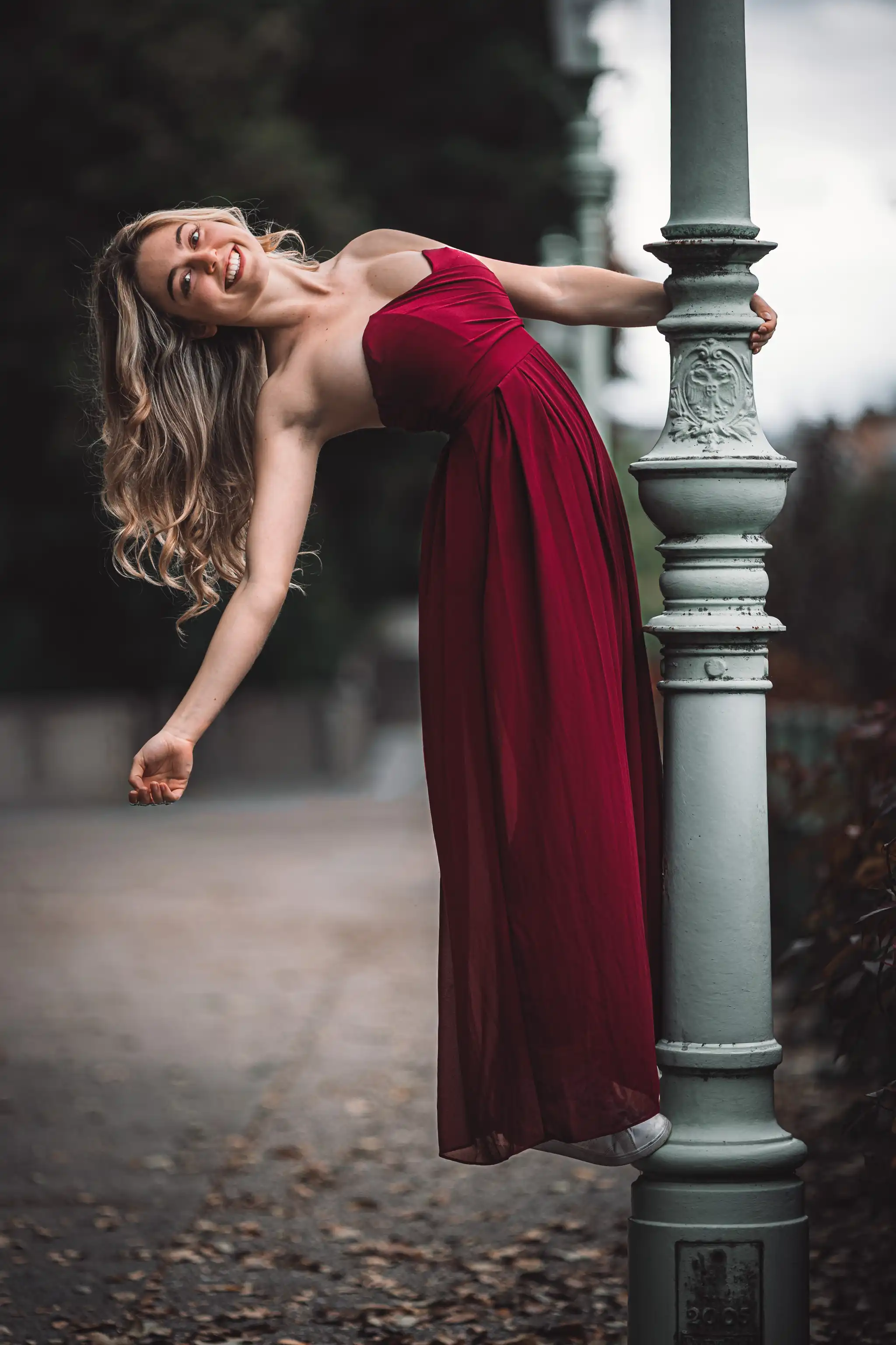 Model Valentina posiert bei einem Porträt-Shooting im Stadtpark Wien in einem eleganten roten Kleid und lehnt sich lachend an einen historischen Laternenmast zurück