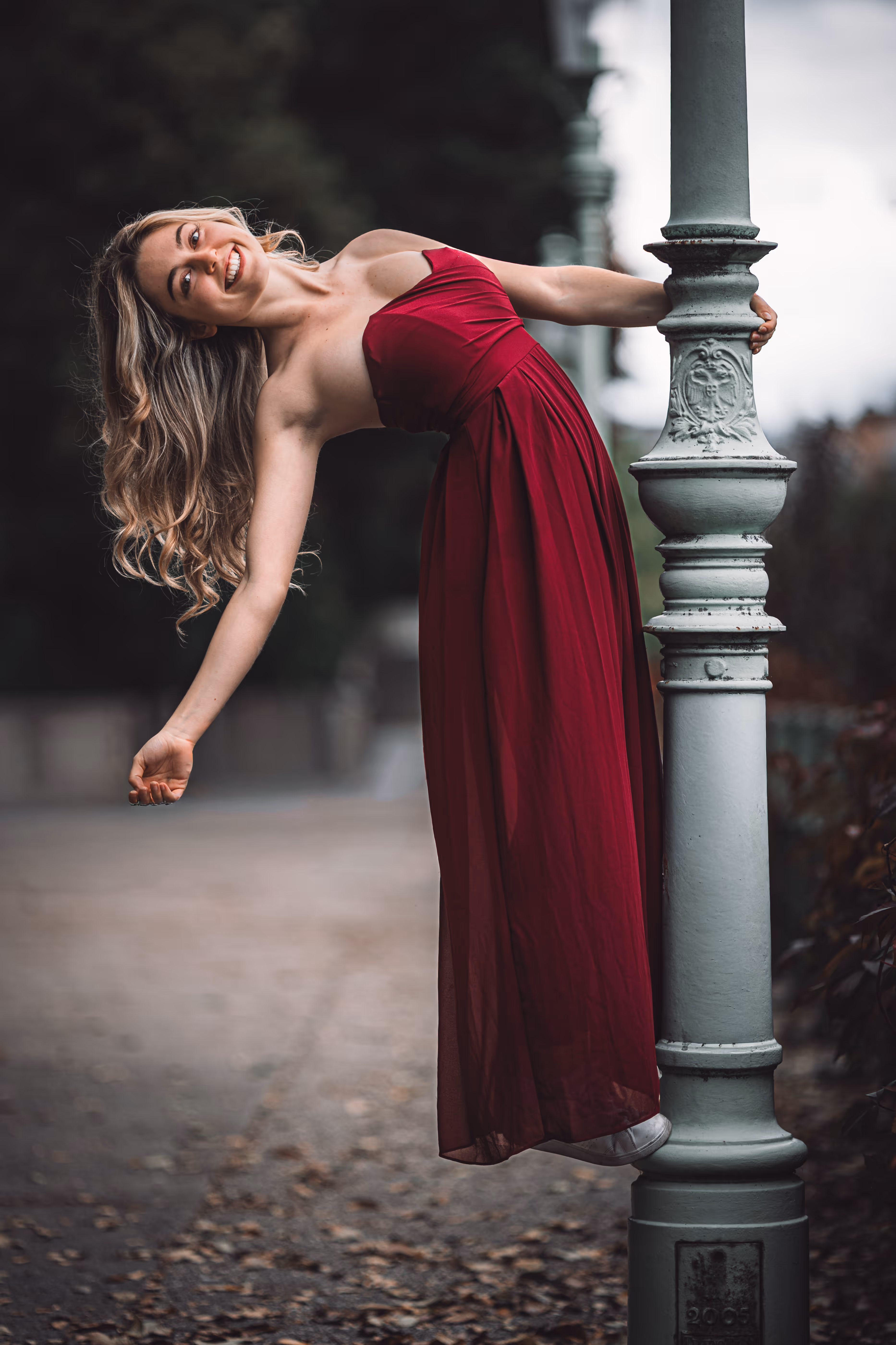 Model Valentina posiert bei einem Porträt-Shooting im Stadtpark Wien in einem eleganten roten Kleid und lehnt sich lachend an einen historischen Laternenmast zurück