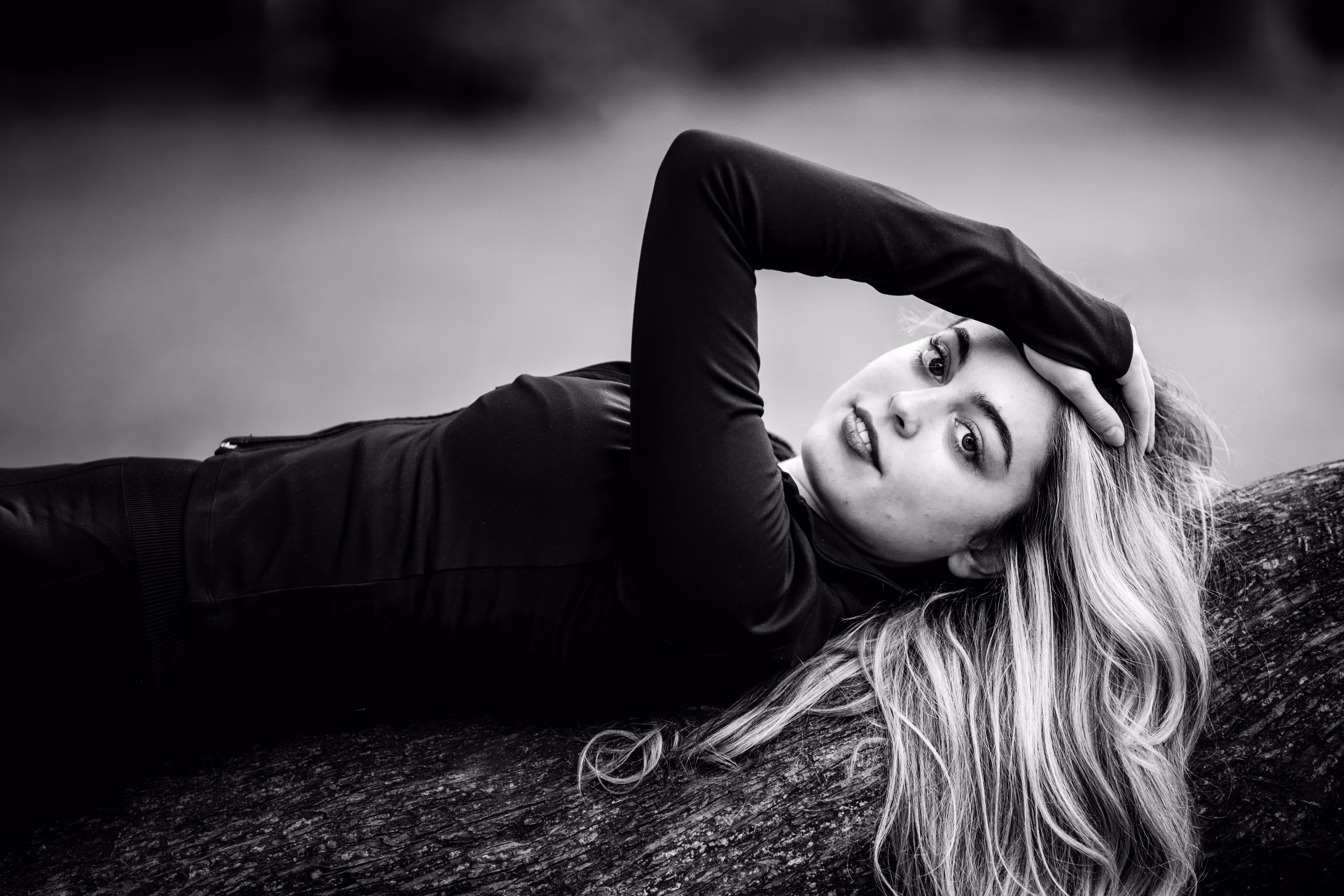 Ausdrucksstarkes Schwarz-Weiß-Porträt von Model Valentina, die auf einem Baumstamm liegt und mit der Hand im Haar direkt in die Kamera blickt