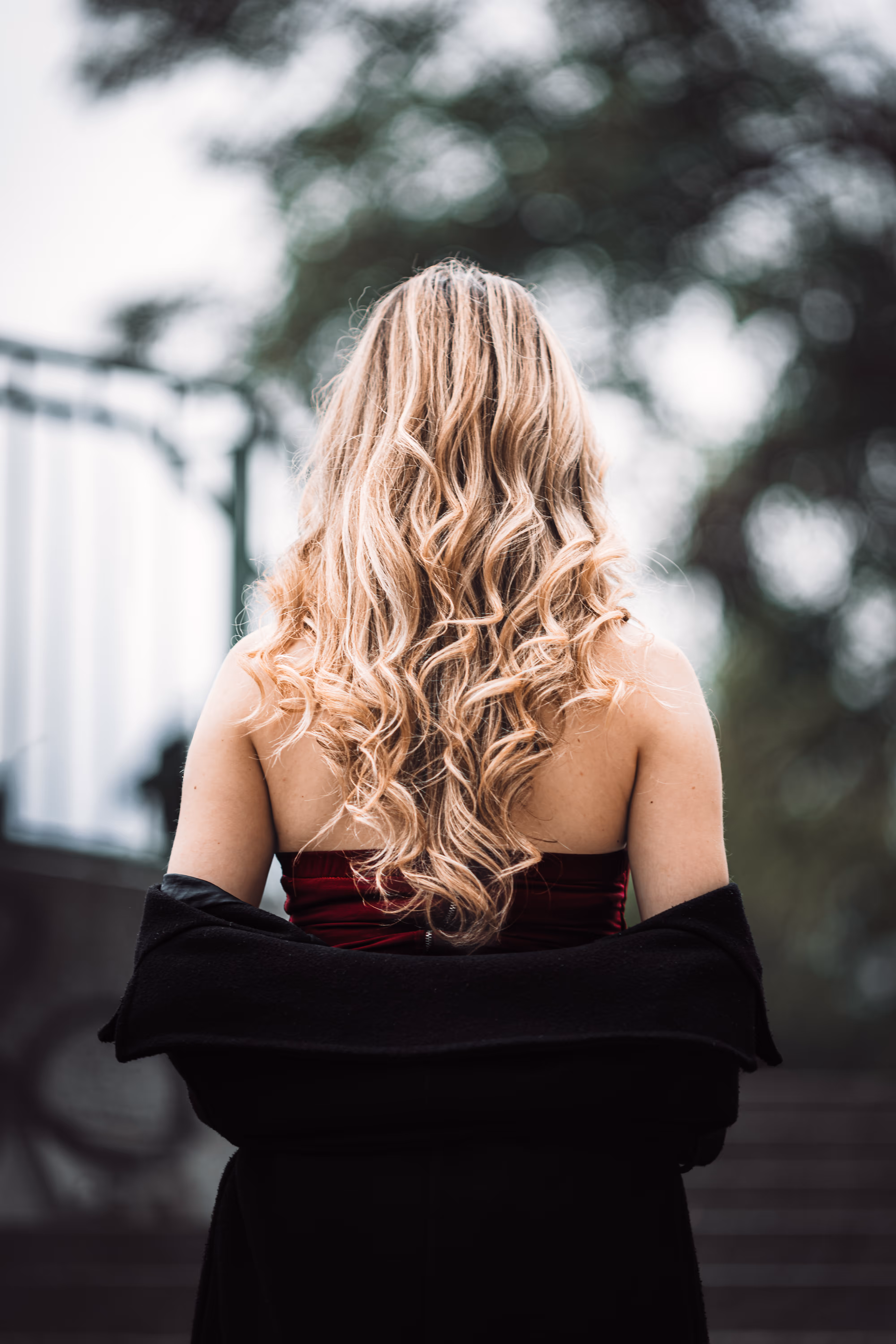 Rückansicht von Model Valentina mit langen, blonden Locken, die während ihres Porträt-Shootings im Wiener Stadtpark ein rotes Kleid und einen schwarzen Mantel trägt