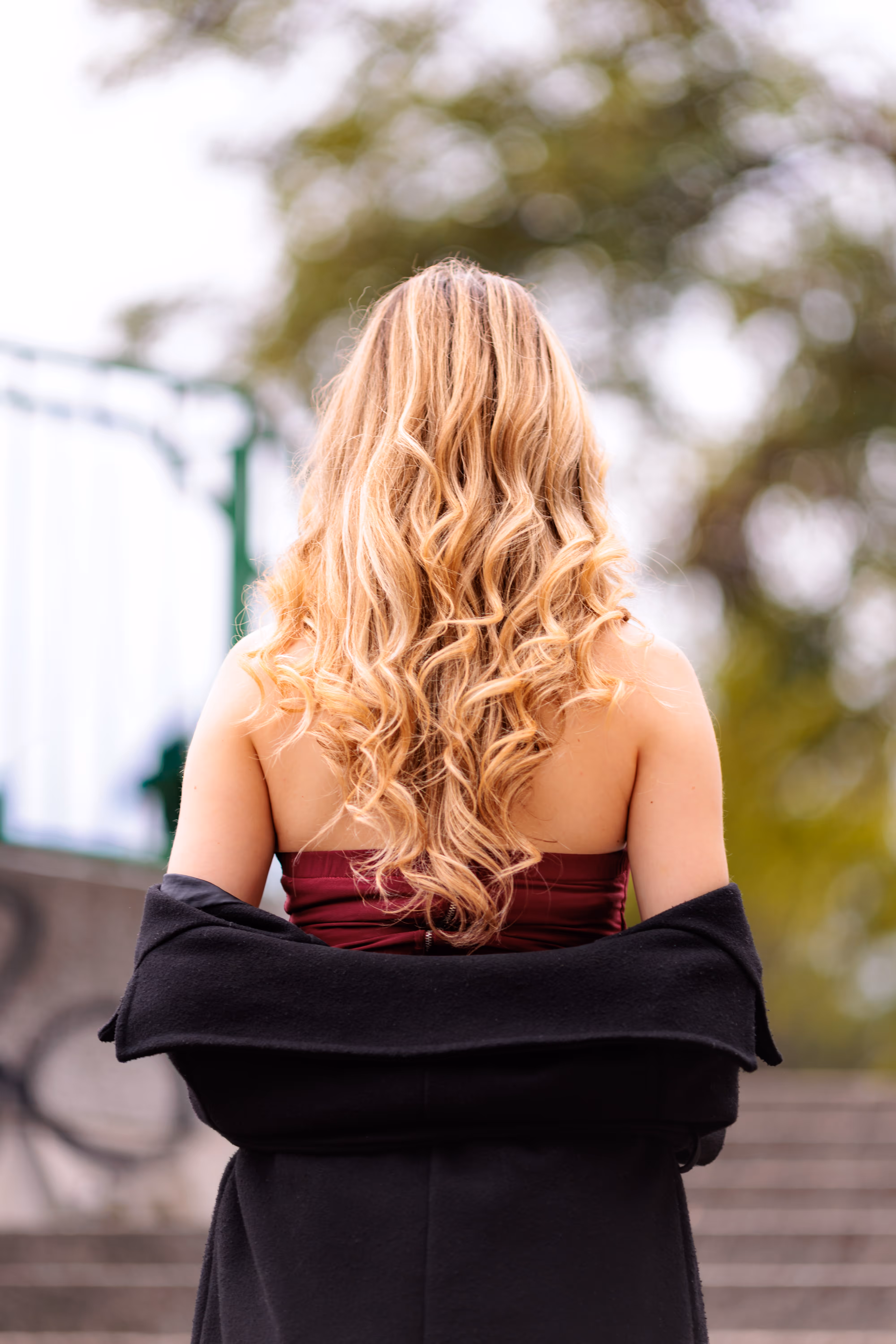 Rückenansicht des Models Valentina mit langen, blonden Locken, die ein rotes, schulterfreies Oberteil und einen lässig über die Schultern gelegten schwarzen Mantel trägt
