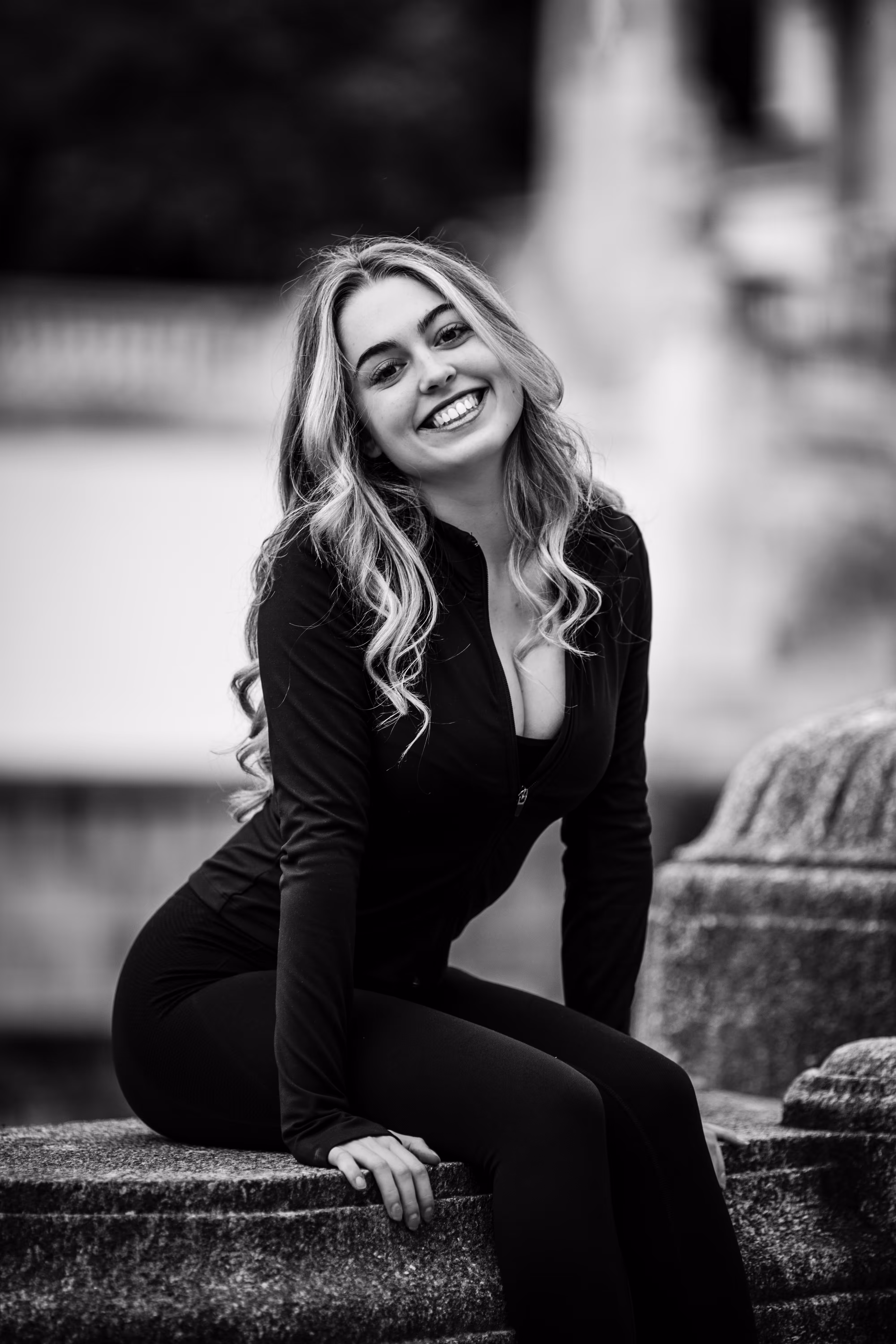 Schwarz-Weiß-Porträt von Model Valentina, die in sportlicher Kleidung auf einer Steinmauer im Stadtpark sitzt und strahlend in die Kamera lächelt