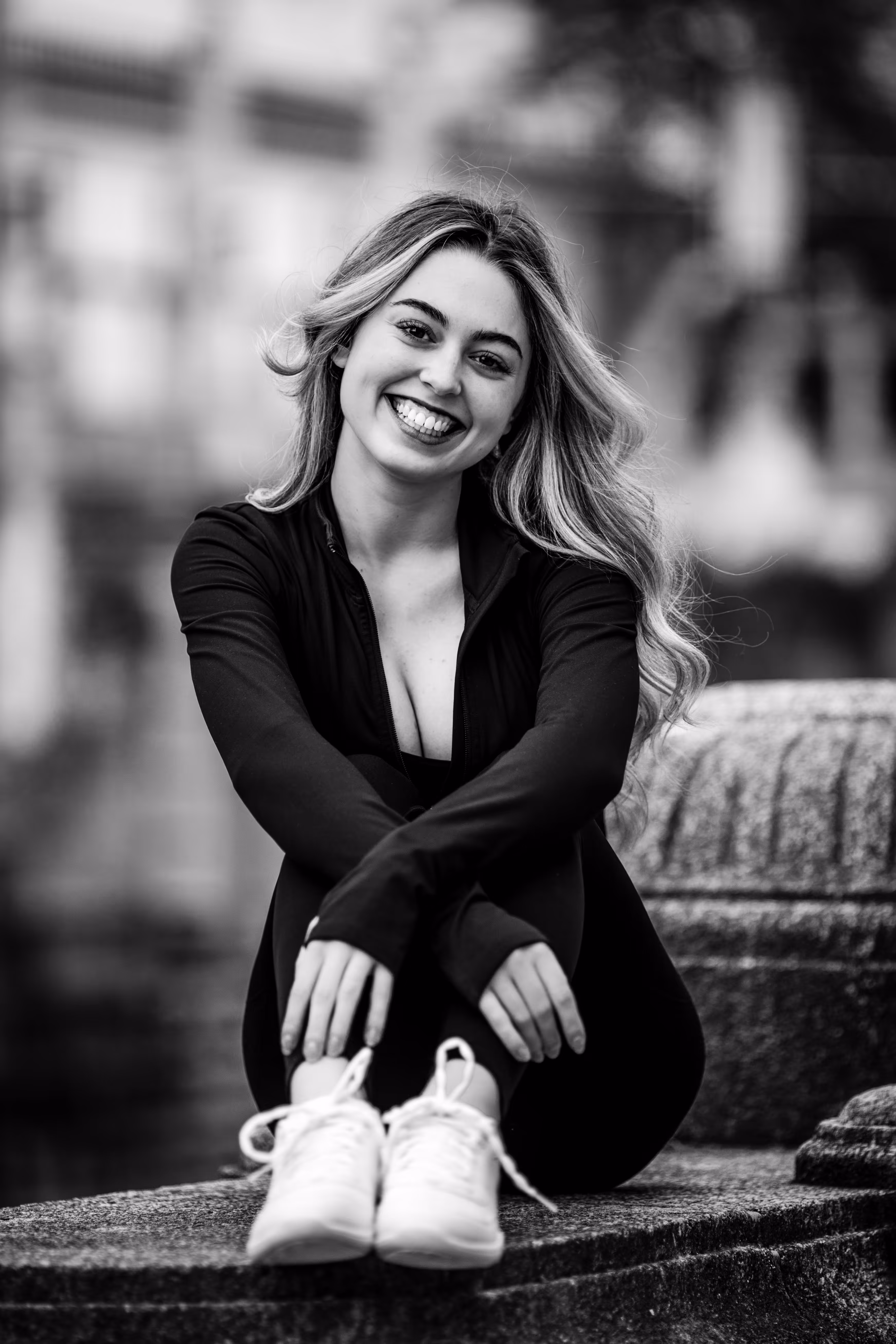 Ausdrucksstarkes Schwarz-Weiß-Porträt von Model Valentina, die in einem dunklen, sportlichen Outfit auf einer Steinmauer im Stadtpark Wien sitzt und herzlich lacht