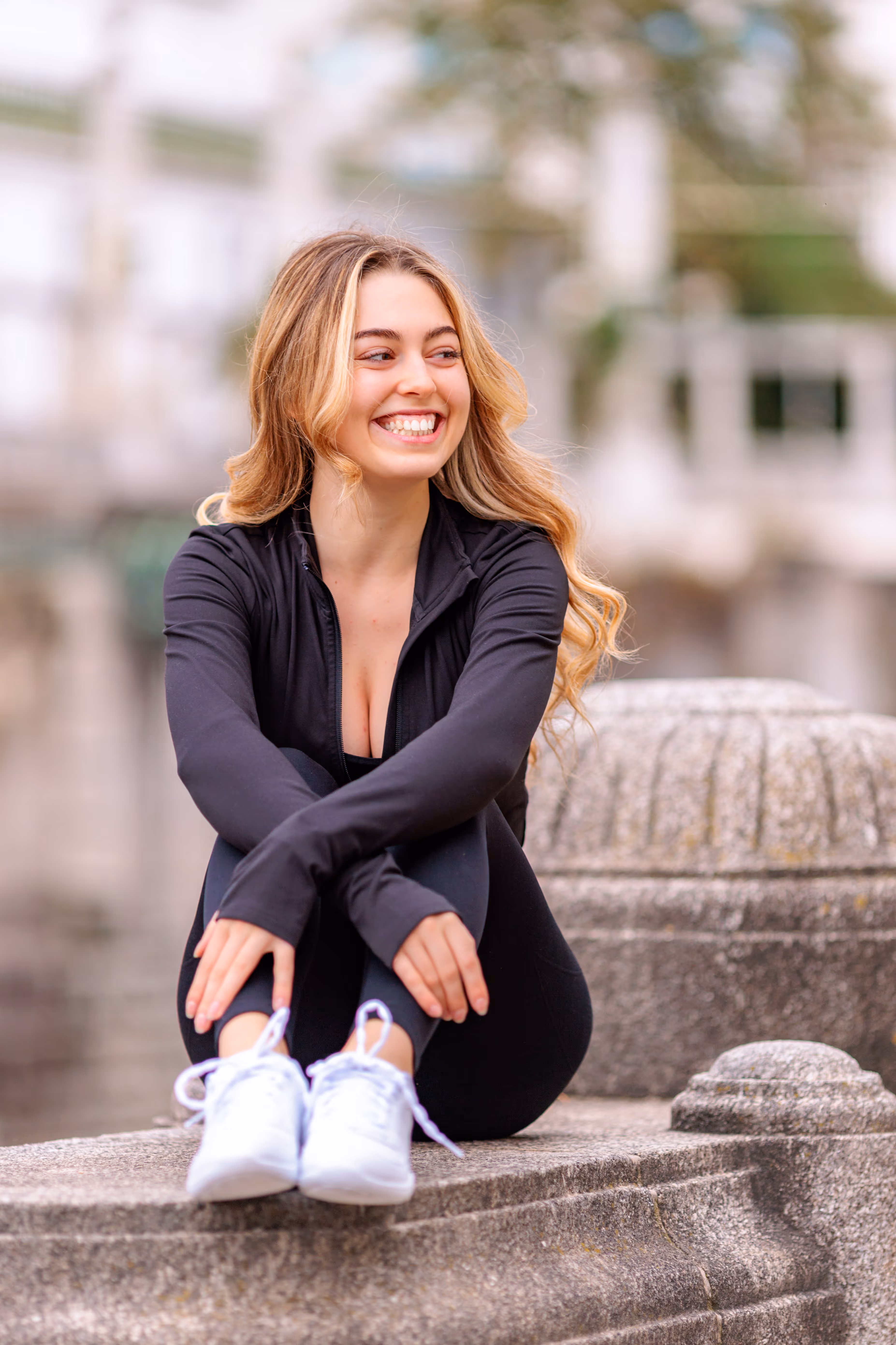 Model Valentina lacht herzlich, während sie in sportlicher schwarzer Kleidung auf einer Steinmauer im Wiener Stadtpark sitzt