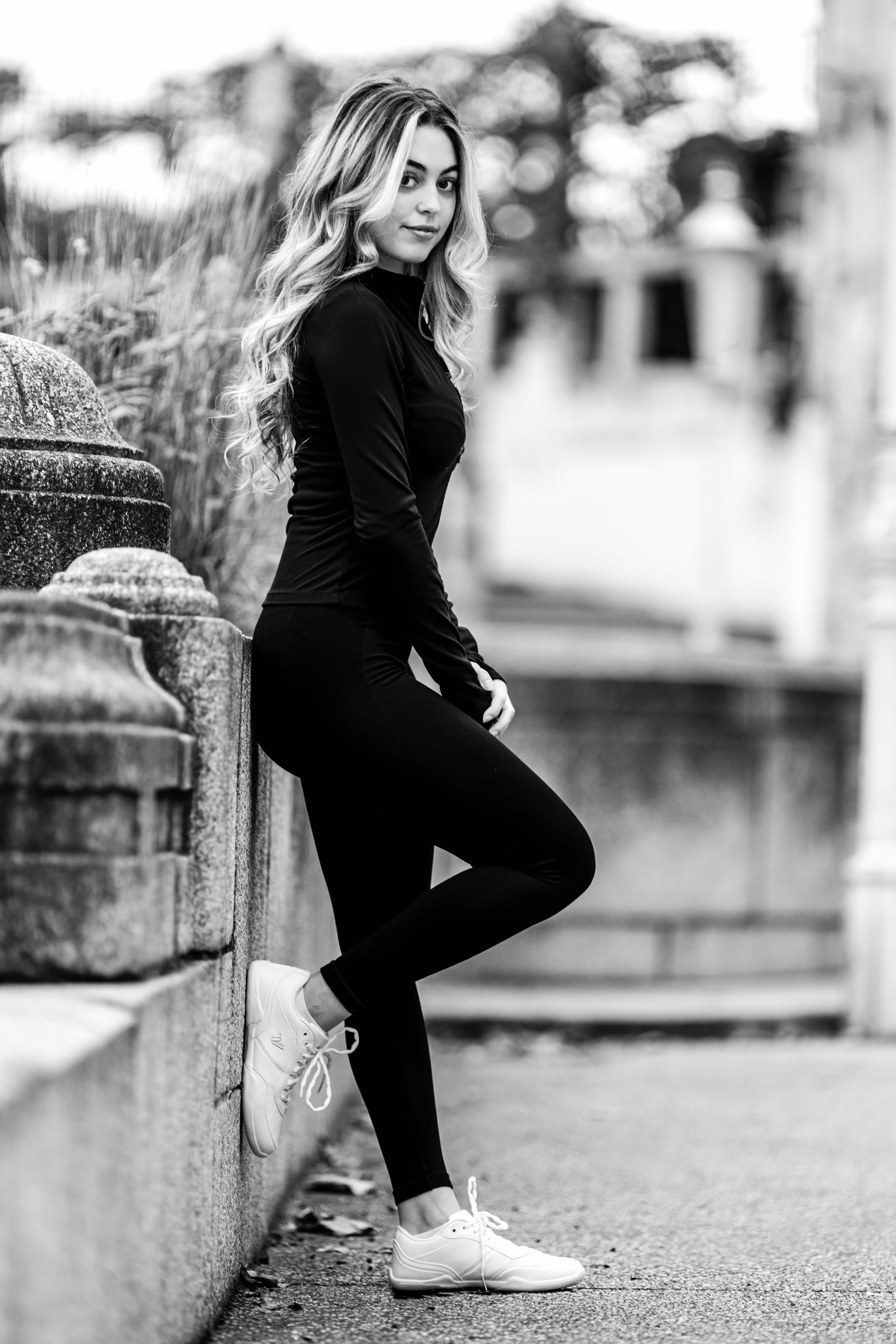Schwarz-Weiß-Ganzkörperporträt von Model Valentina, die in einem sportlichen schwarzen Outfit an einer Steinmauer im Stadtpark lehnt und in die Kamera blickt