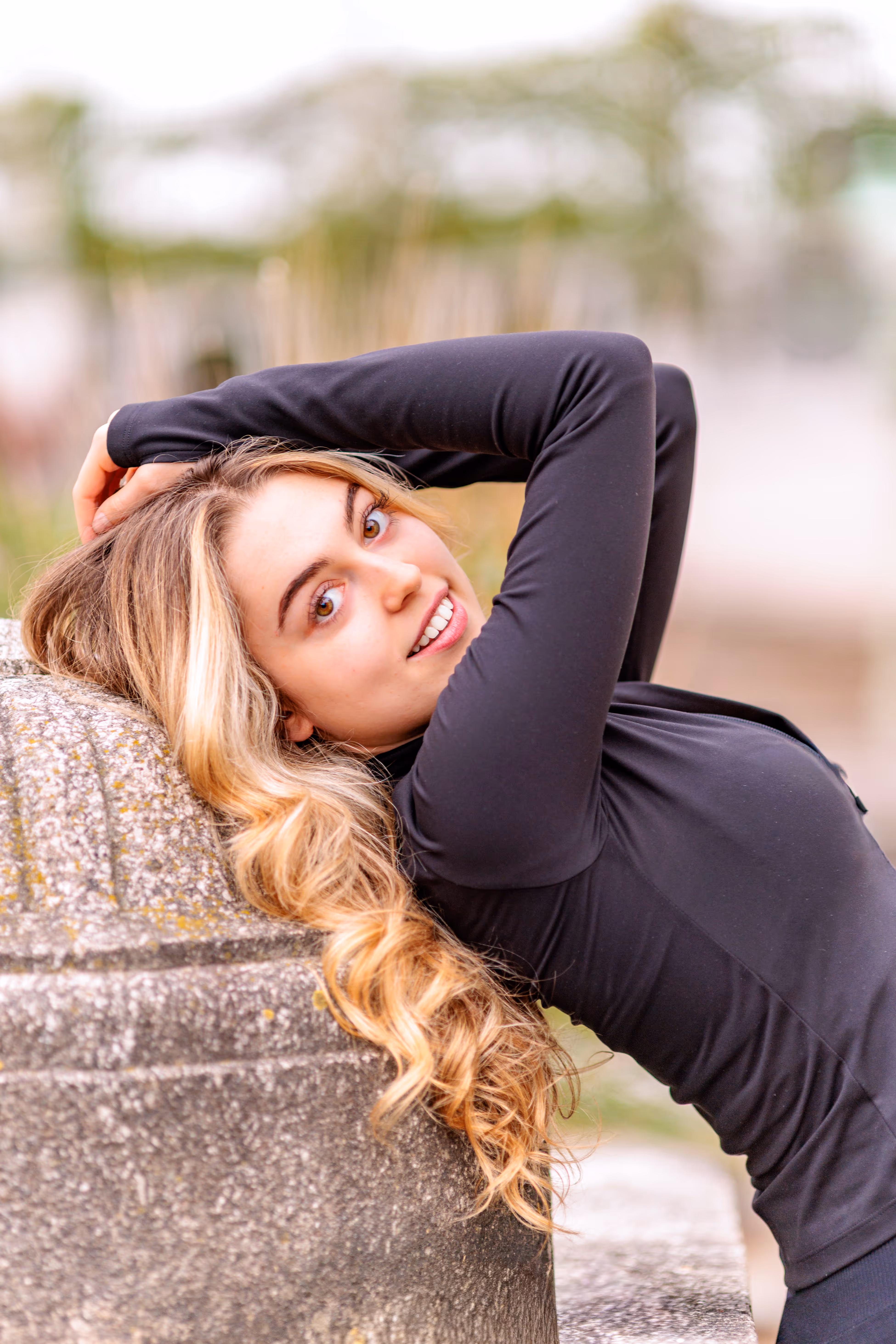 Das junge Model Valentina lehnt sich mit langen, blonden Haaren an eine Steinsäule, hat die Hände hinter dem Kopf und blickt lächelnd in die Kamera
