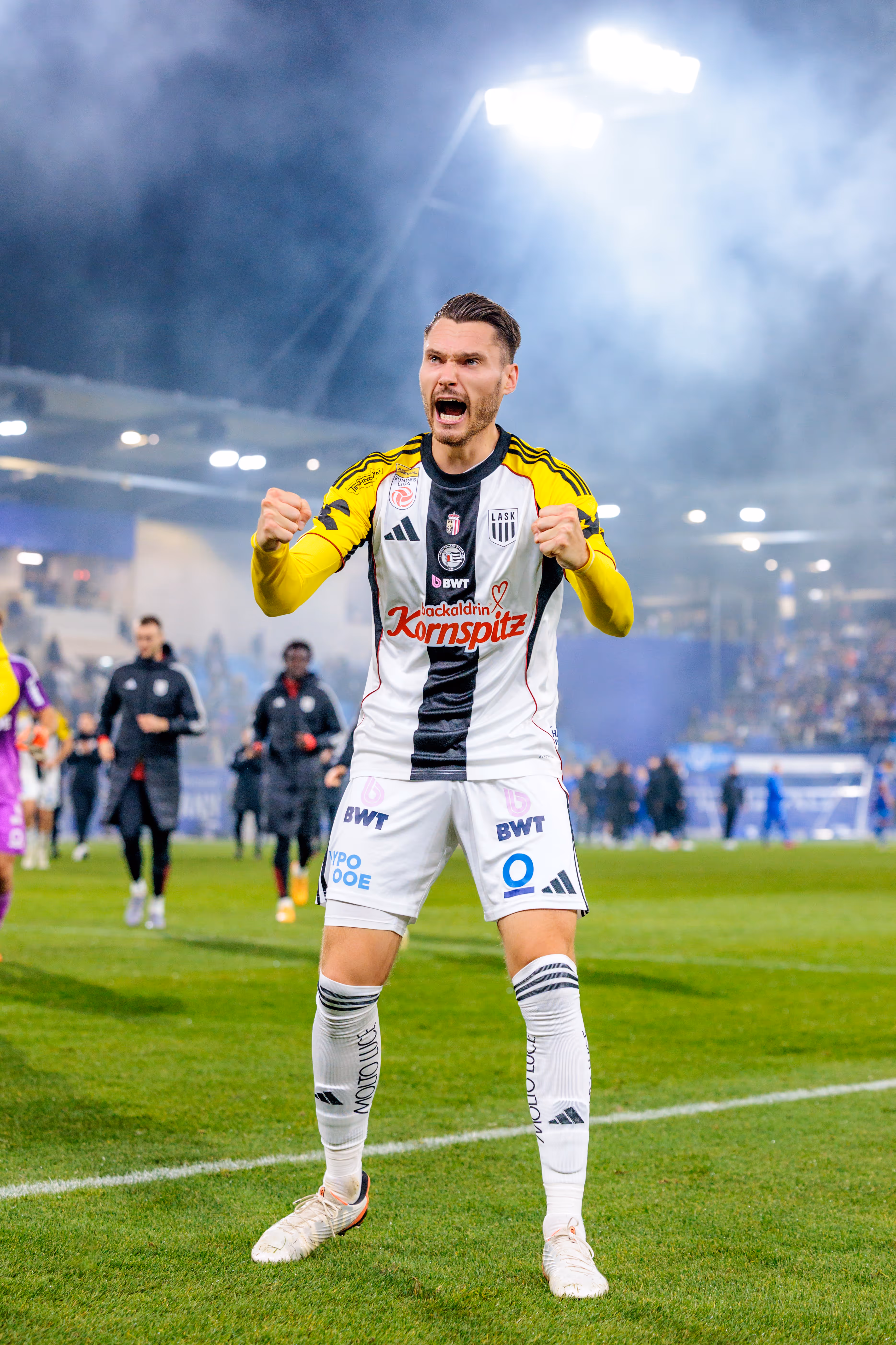 Der LASK-Spieler Valon Berisha ballt in einer kraftvollen Jubelpose die Fäuste und schreit seine Freude heraus, während im Hintergrund des Stadions dichter Rauch aufsteigt