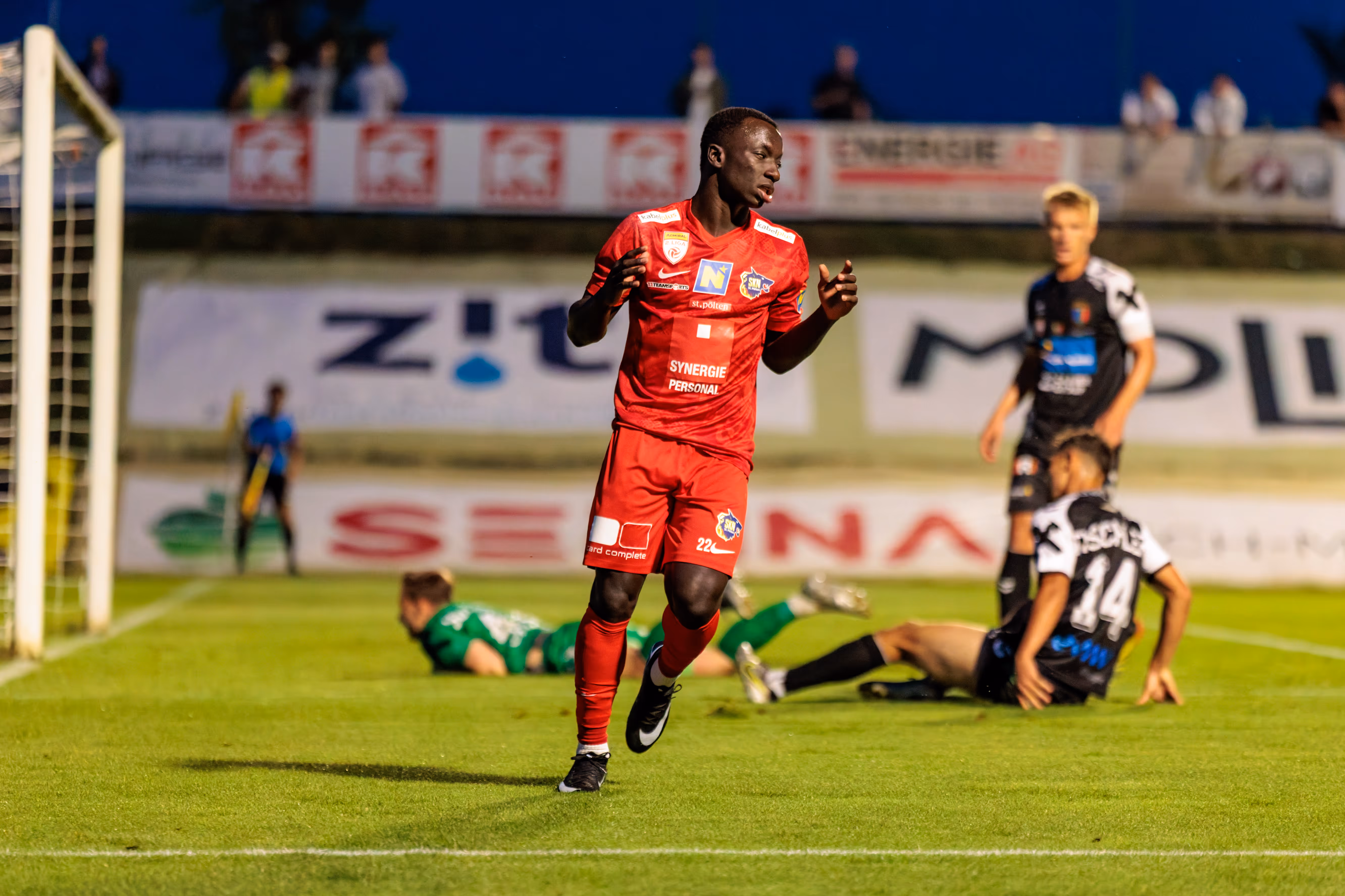 Ein Fußballspieler des SKN St. Pölten in rotem Trikot steht nach einer verpassten Torchance auf dem Spielfeld, während weitere Akteure und Werbebanden im Hintergrund unscharf zu sehen sind