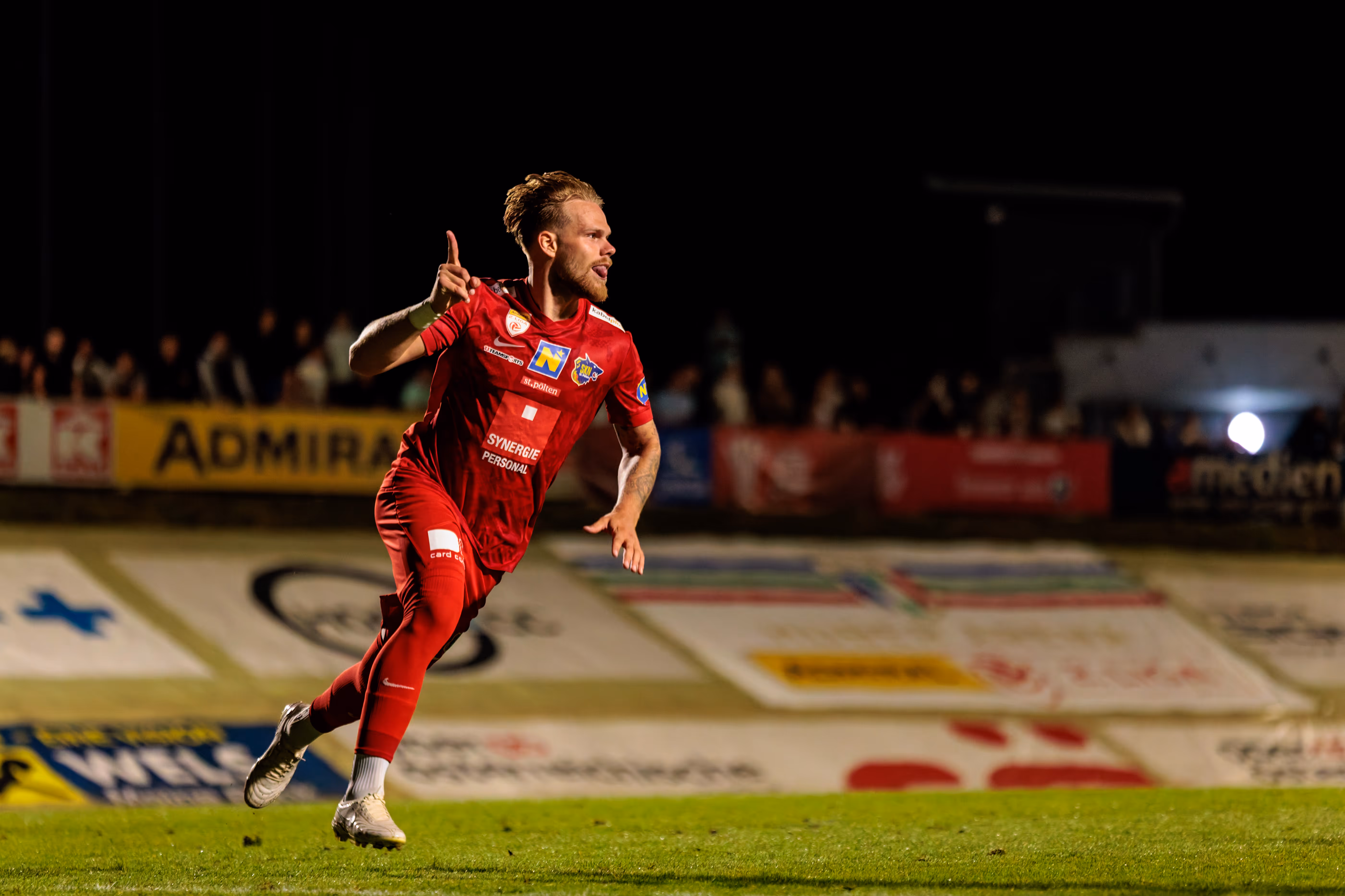 Ein Fußballspieler des SKN St. Pölten in roter Spielkleidung feiert auf dem nächtlichen Spielfeld ausgelassen einen Erfolg mit erhobenem Finger