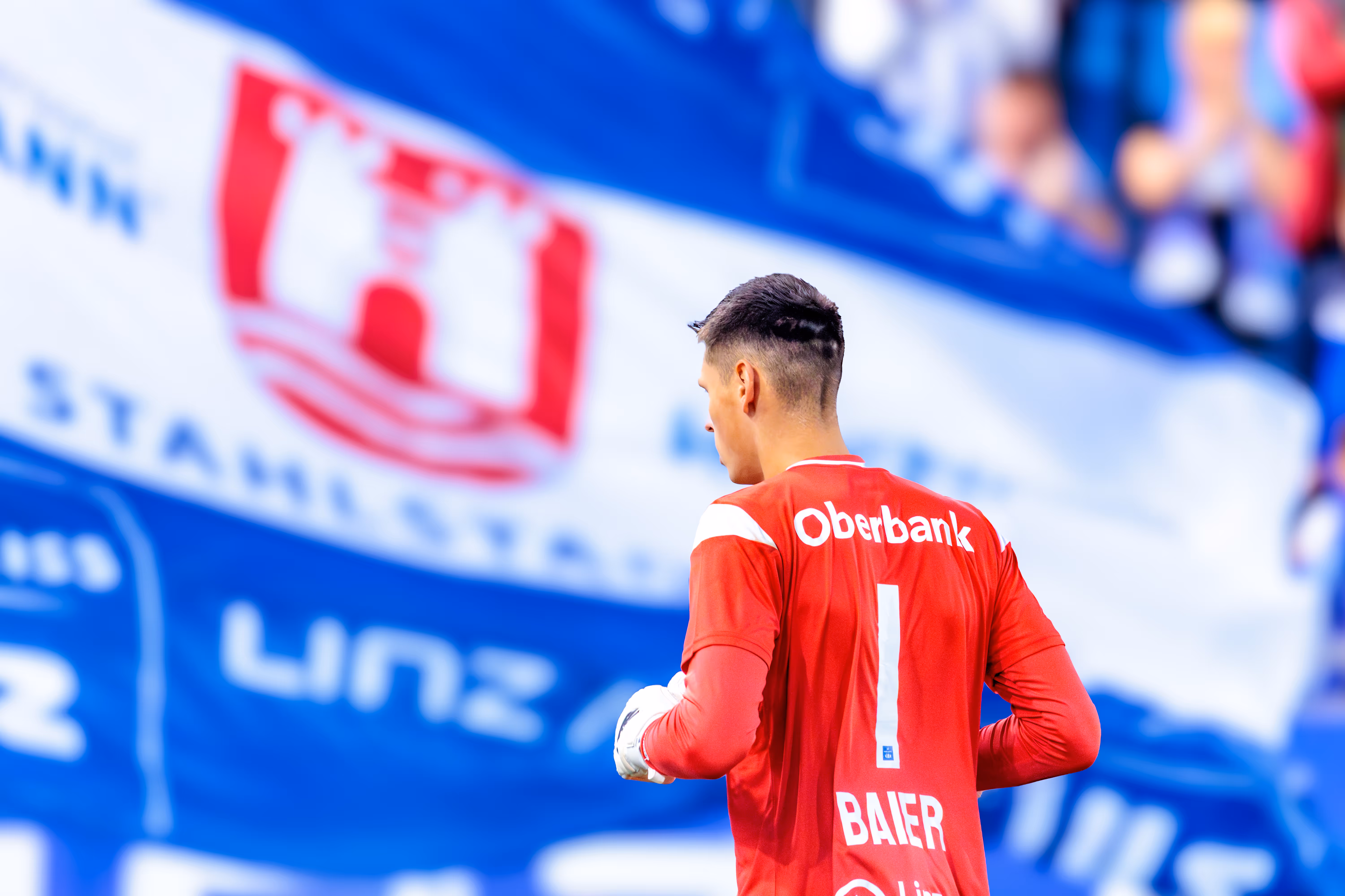 Rückansicht des Torhüters Viktor Baier vom FC Blau-Weiß Linz in seinem roten Trikot mit der Nummer 1 vor einer großen, unscharfen Fan-Fahne im Hintergrund