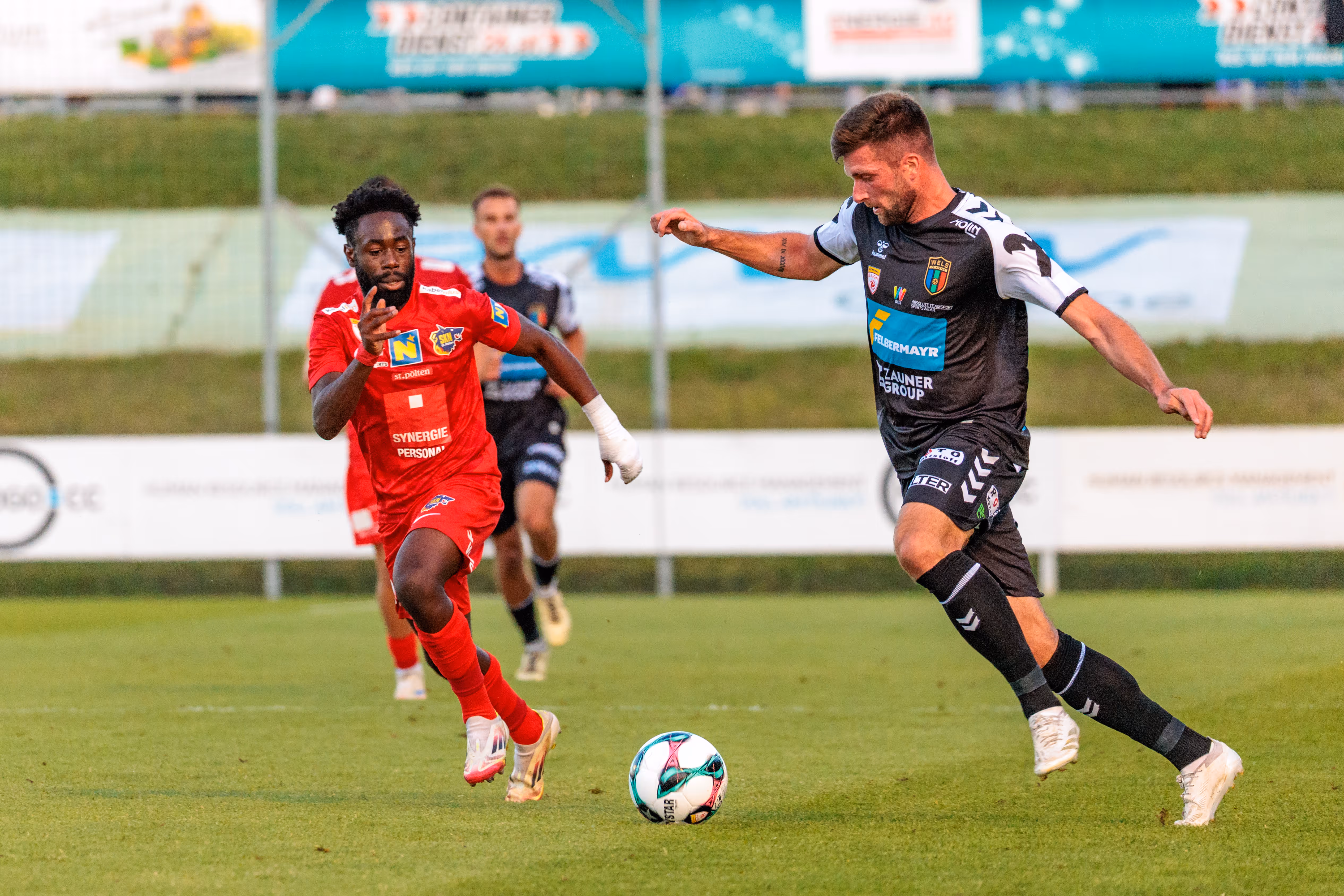 Zwei Fußballer, ein Spieler des SKN St. Pölten in rotem Trikot und ein Spieler der SPG HOGO Wels in schwarz-weißem Trikot, streiten im ÖFB-Cup-Match um den Ball