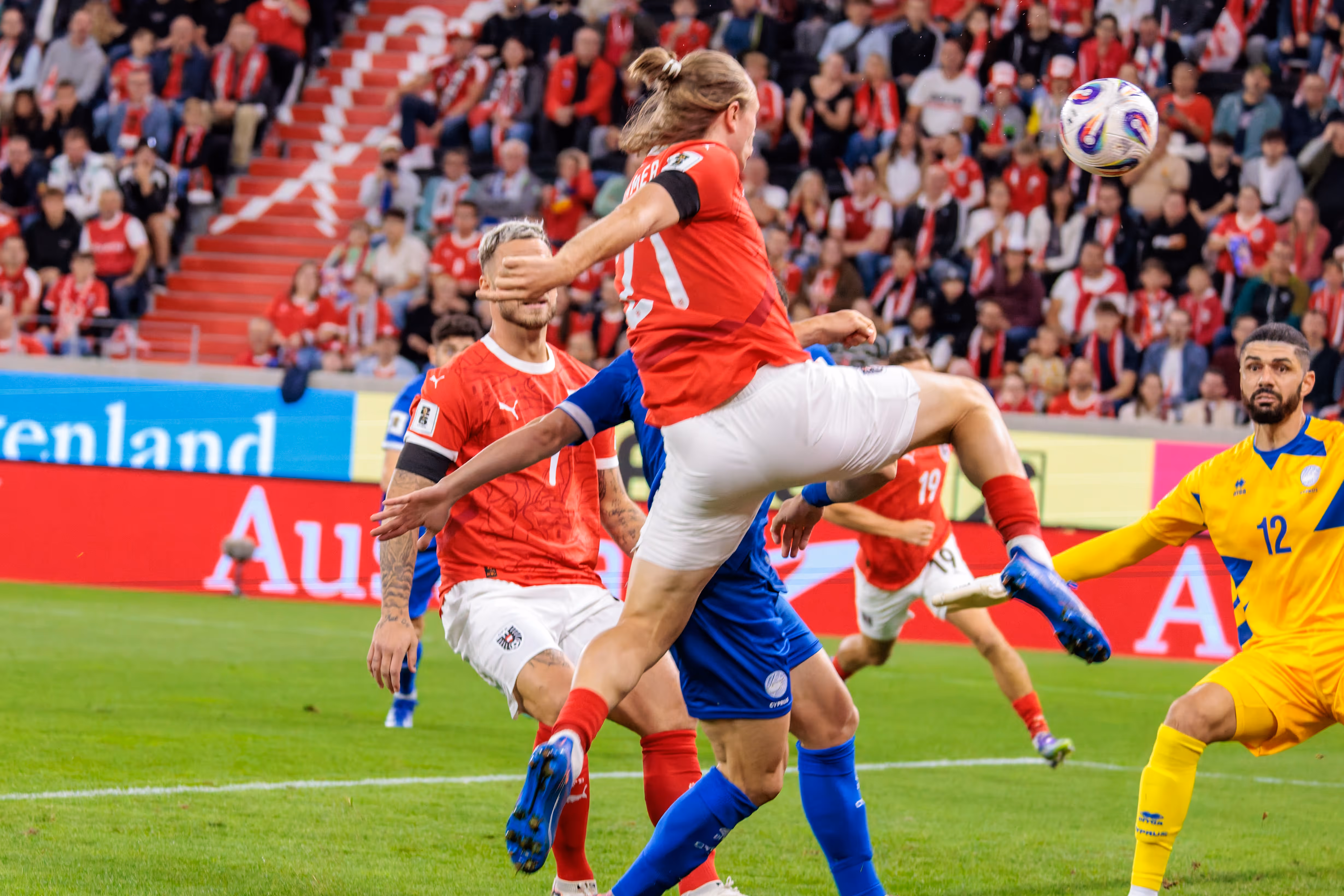 Ein österreichischer Spieler mit der Rückennummer 21, Patrick Wimmer, steigt im Strafraum zum Ball hoch, während der zypriotische Torhüter Fabiano mit der Nummer 12 das Spiel aufmerksam verfolgt