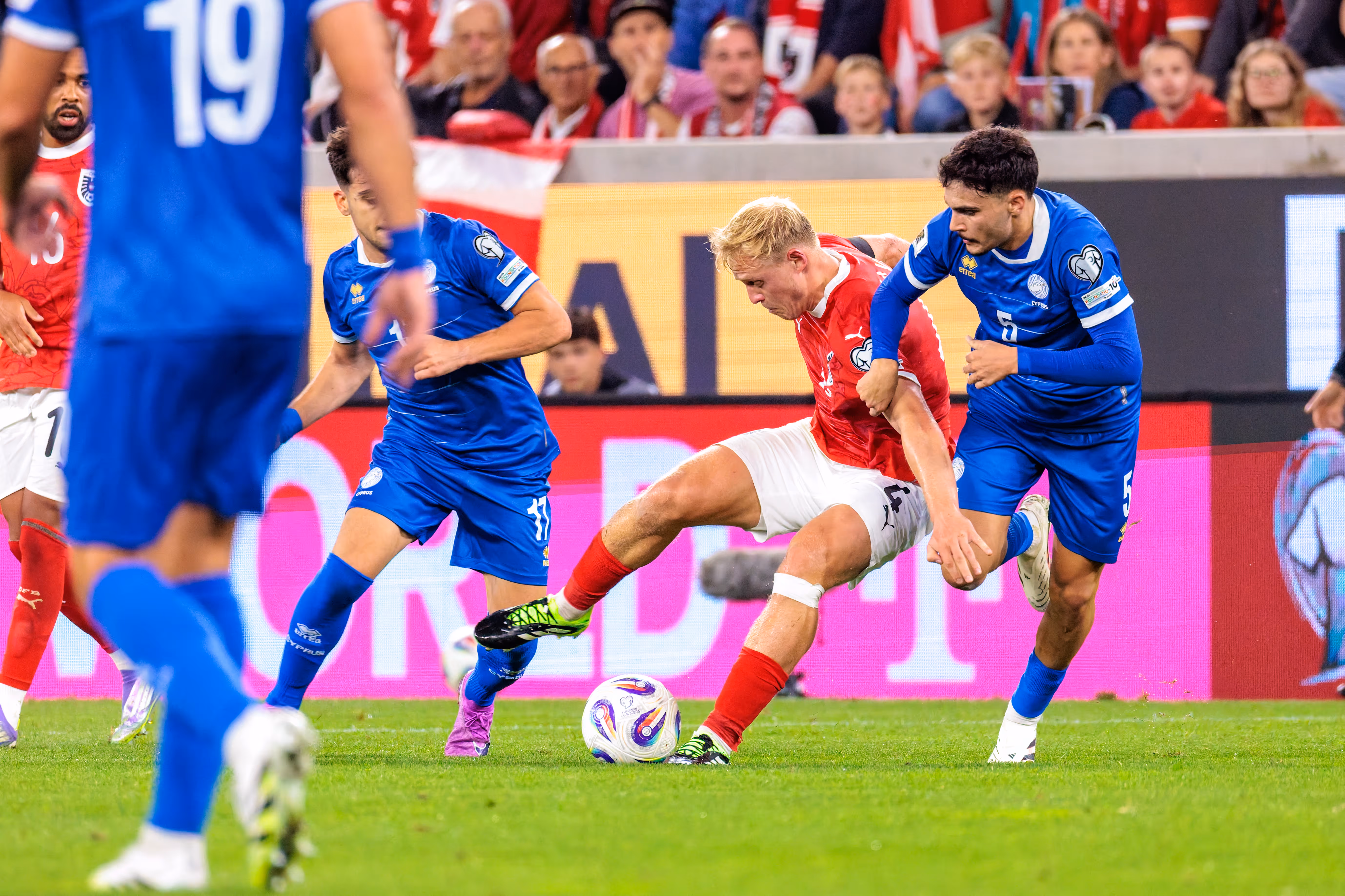 Xaver Schlager von Österreich im roten Trikot ringt im Zweikampf mit Charalampos Charalampous von Zypern im blauen Trikot um den Ball, während Konstantinos Laifis im Vordergrund das geschehen beobachtet